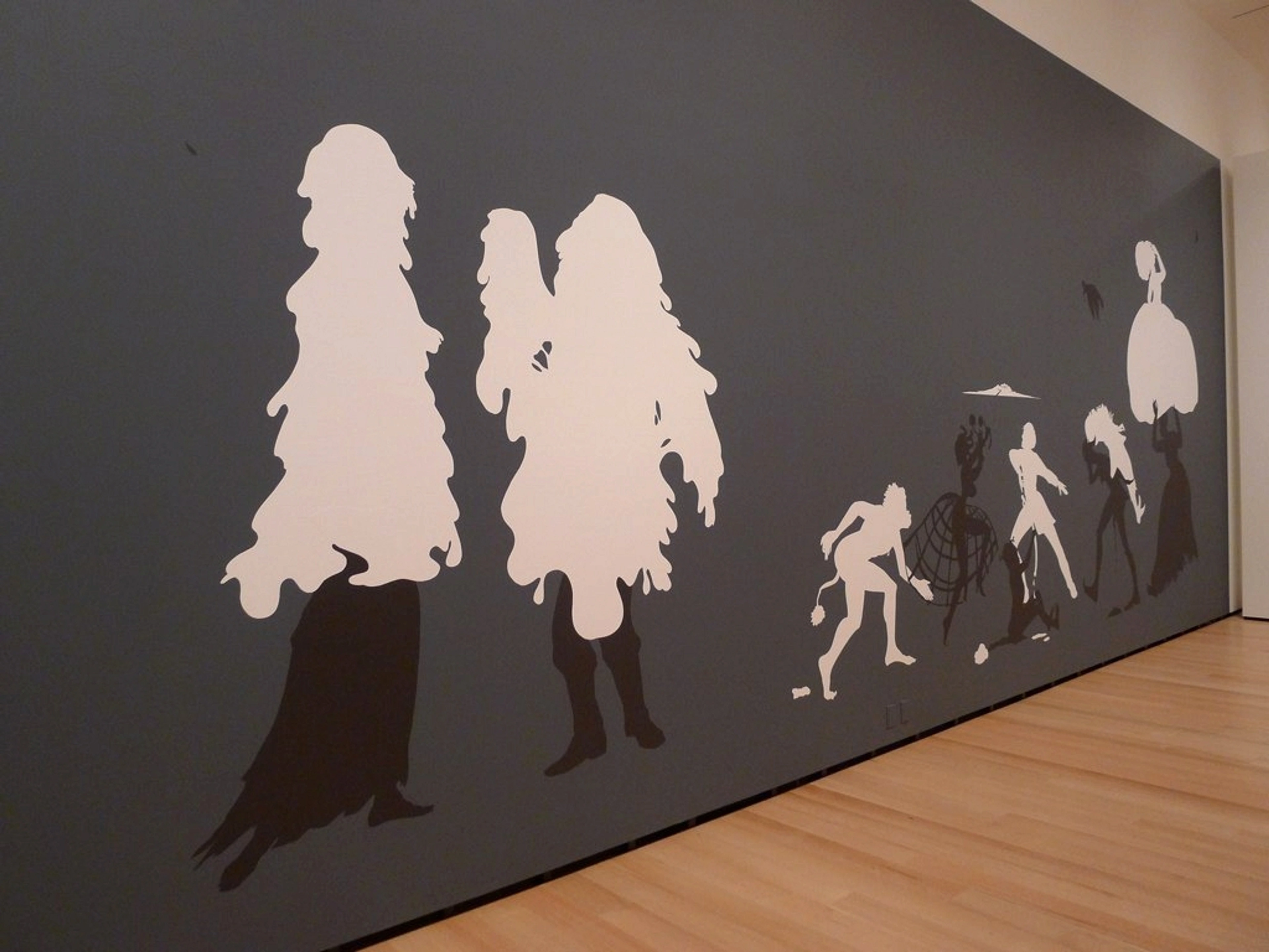 Kara Walker's 'The Rich Soil Down There' mural, featuring large white and smaller black and white silhouettes of figures on a dark grey wall in a museum setting.
