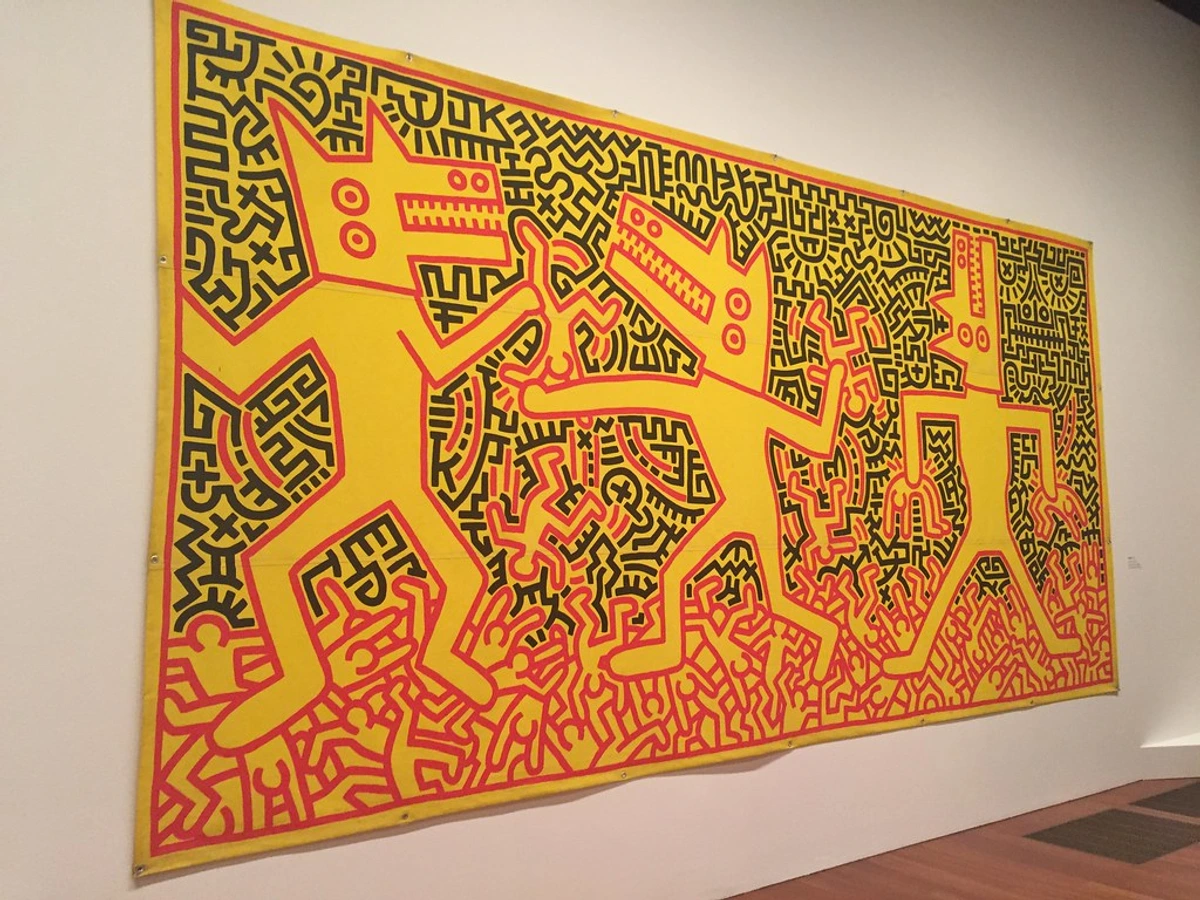 Large Keith Haring artwork featuring three yellow dog-like figures with black outlines and red details, set against a background of black and red abstract patterns, displayed on a white wall in a museum.