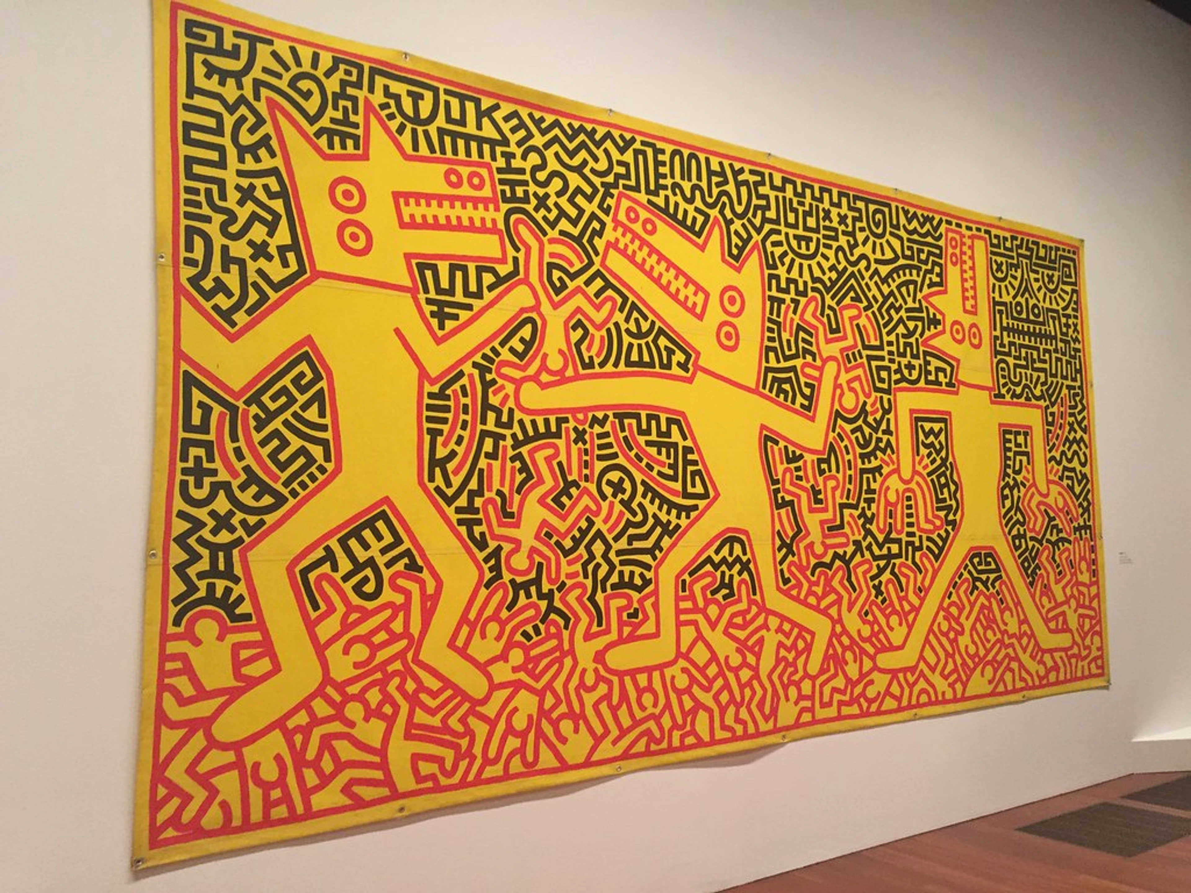 Large Keith Haring artwork featuring three yellow dog-like figures with black outlines and red details, set against a background of black and red abstract patterns, displayed on a white wall in a museum.