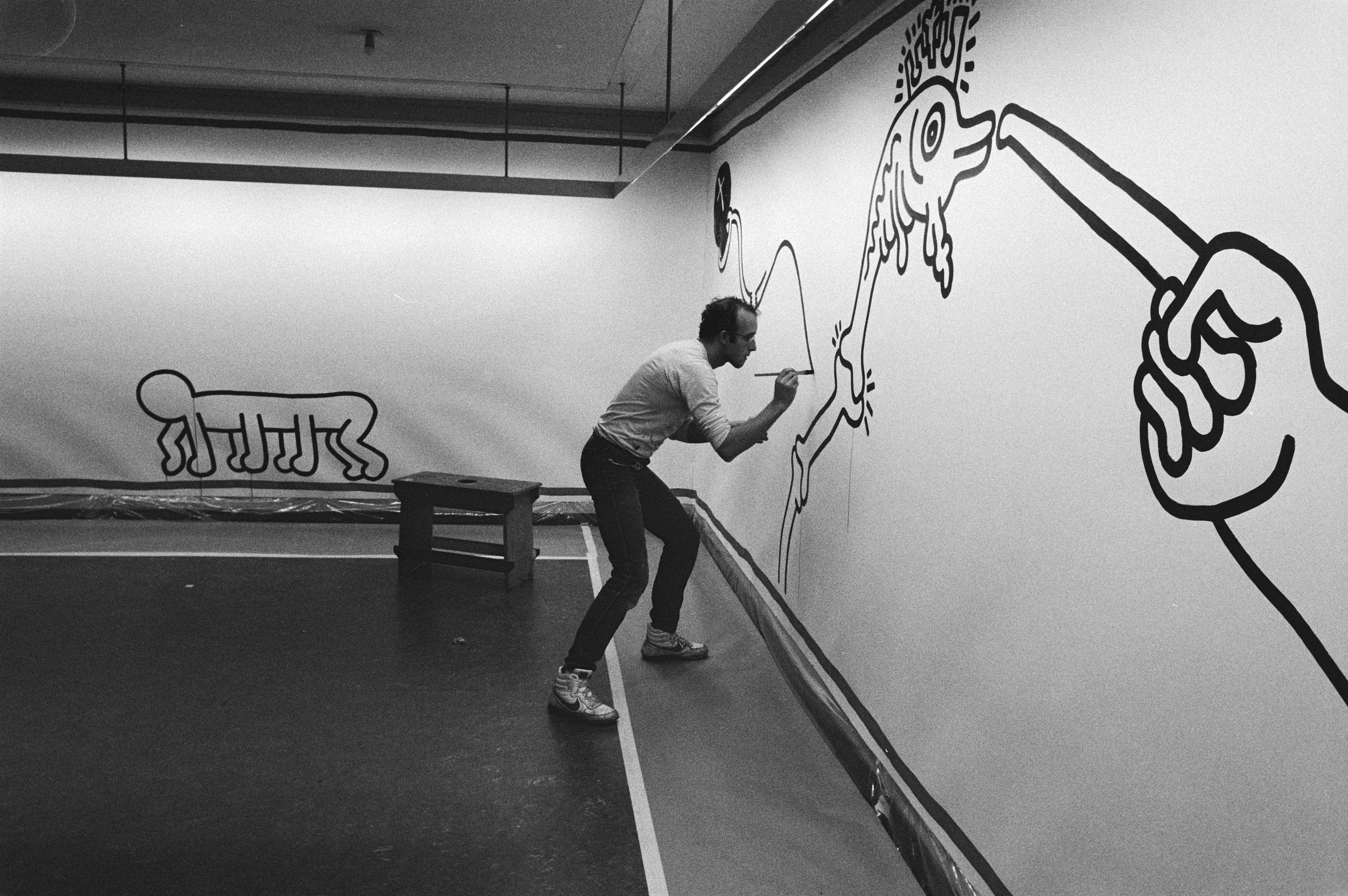 Keith Haring painting a large black line art mural in 1986, featuring his iconic figures like a crawling baby and a fish.