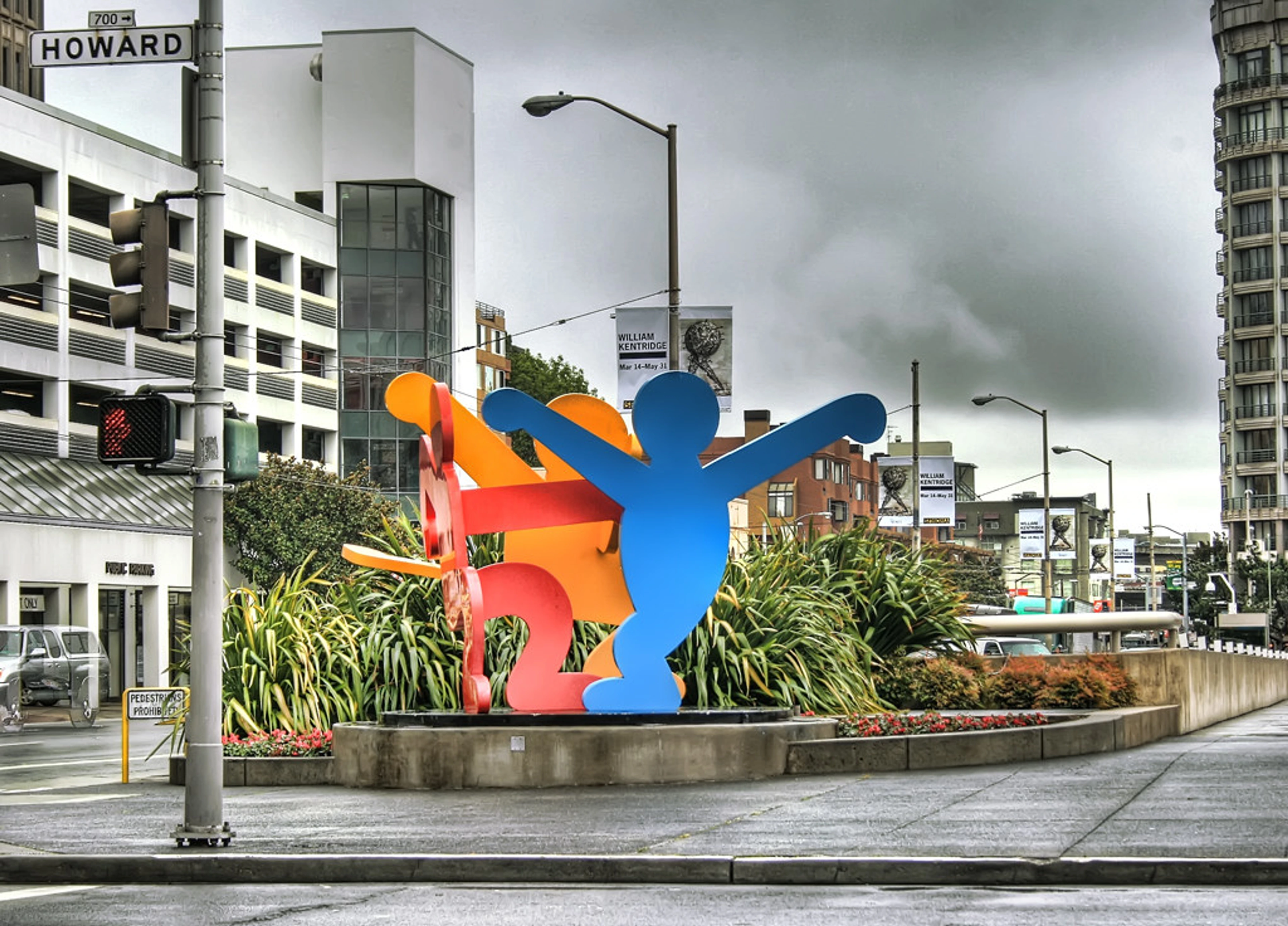 Keith Haring's 'Three Dancing Figures' sculpture, a vibrant blue, orange, and red abstract artwork, stands on a city street corner with buildings and a 'Howard' street sign under an overcast sky.