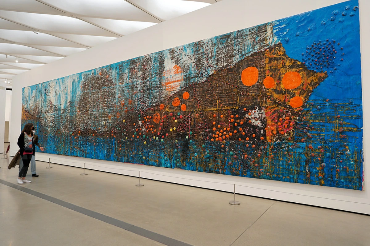 A large, textured abstract painting by Mark Bradford titled 'Deep Blue' in a modern art gallery, featuring blue, brown, and orange colors with spherical elements, viewed by visitors.