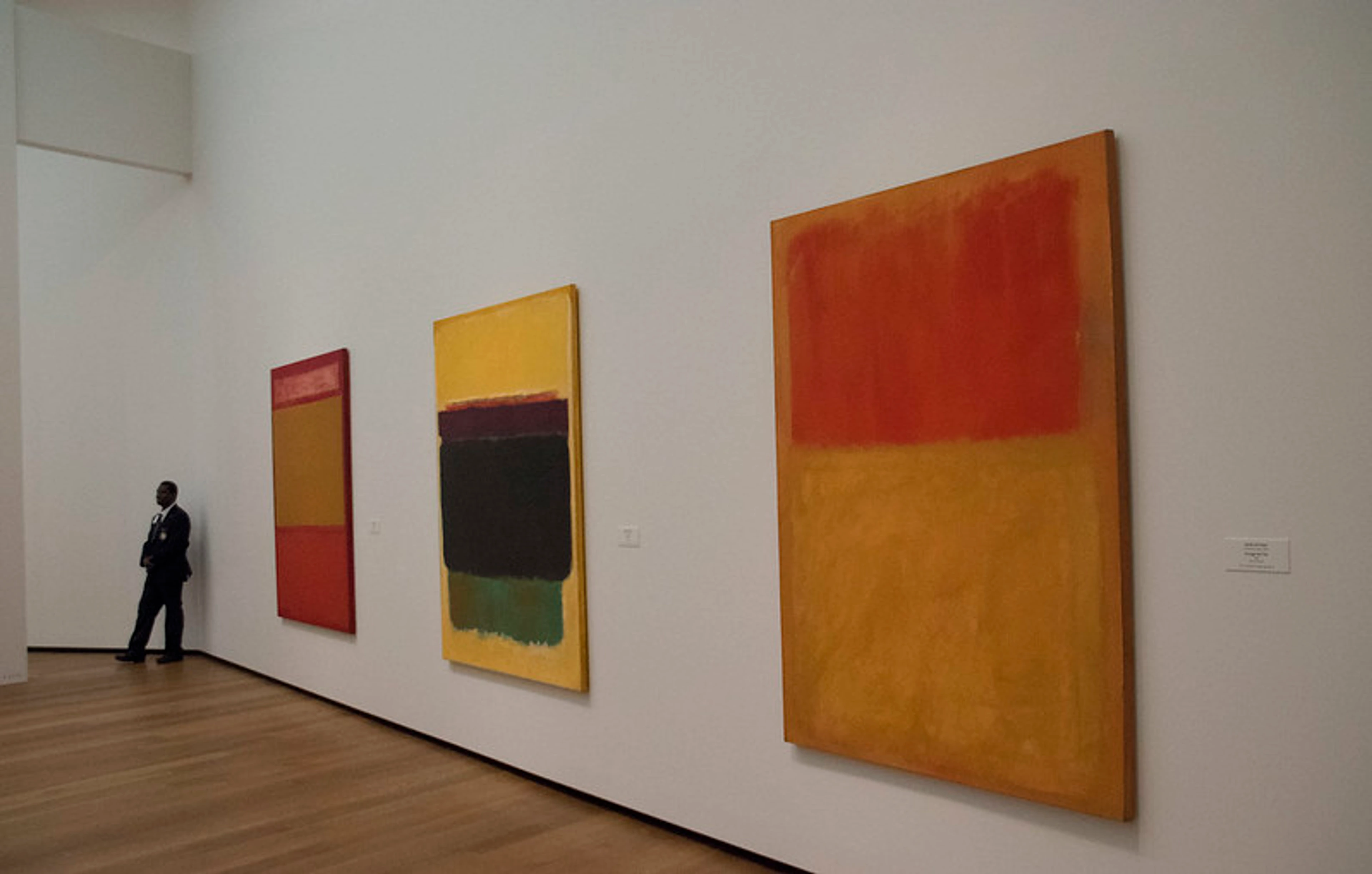 Uitzicht op drie grootschalige kleurvlak schilderijen van Mark Rothko tentoongesteld op een witte muur in het Fondation Beyeler museum, met een bezoeker links staand.
