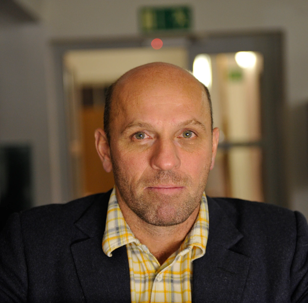 Close-up portrait of artist Peter Doig, a bald man with a beard, wearing a plaid shirt and dark jacket, looking directly at the camera.