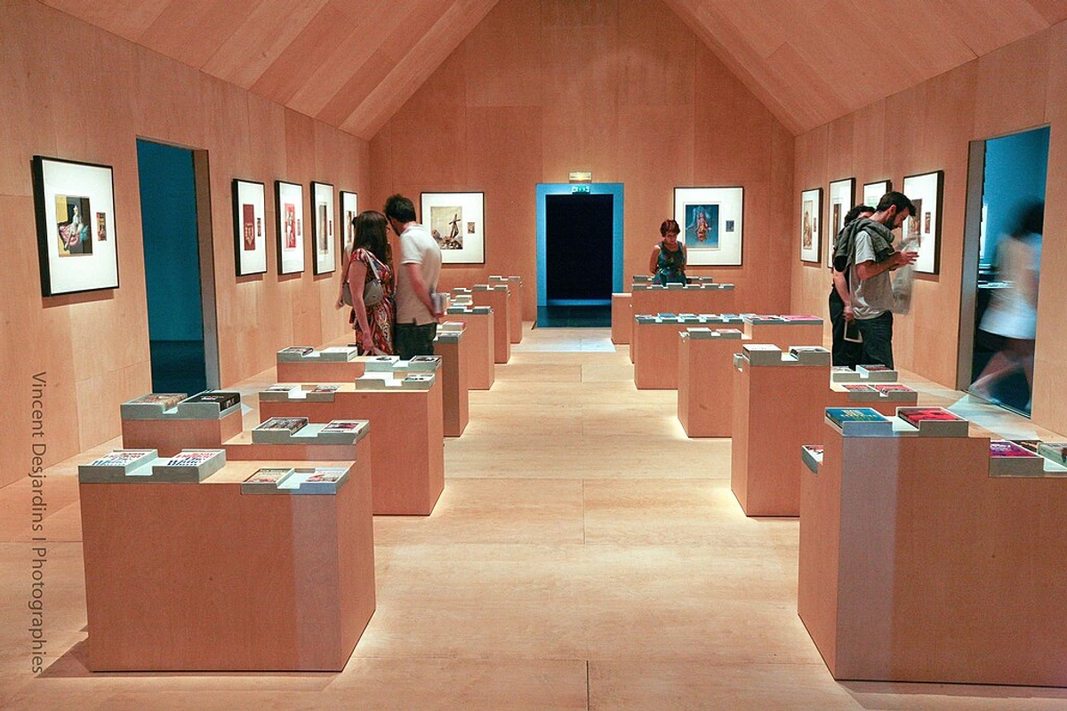 Interior view of an art exhibition with light wooden walls, framed artworks, and display pedestals with books or catalogs, with visitors browsing.