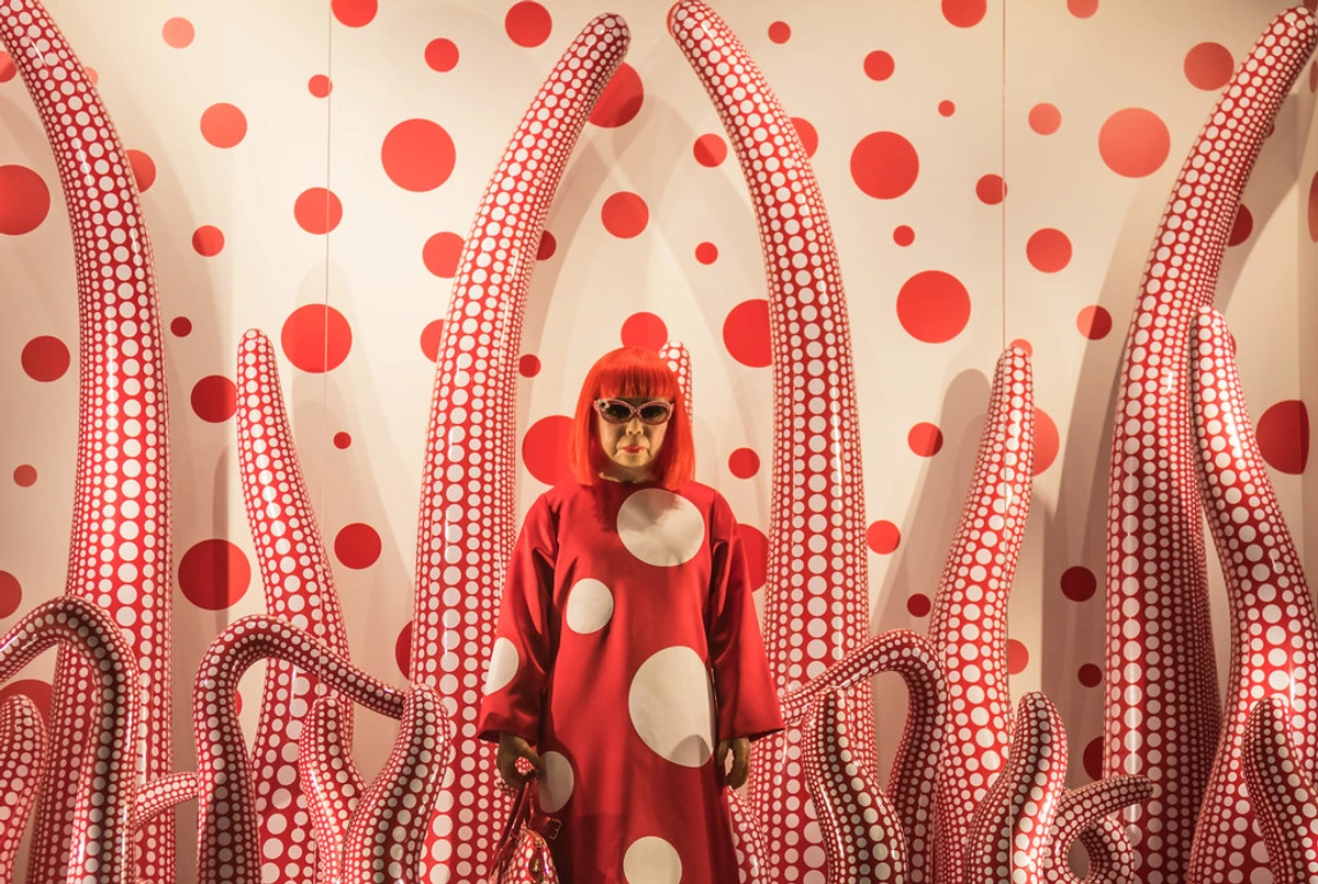 Yayoi Kusama, een Japanse kunstenares, staat in een rood gestippelde jurk en zonnebril te midden van haar iconische rood-witte gestippelde tentakelsulpturen en een bijpassende gestippelde muur.