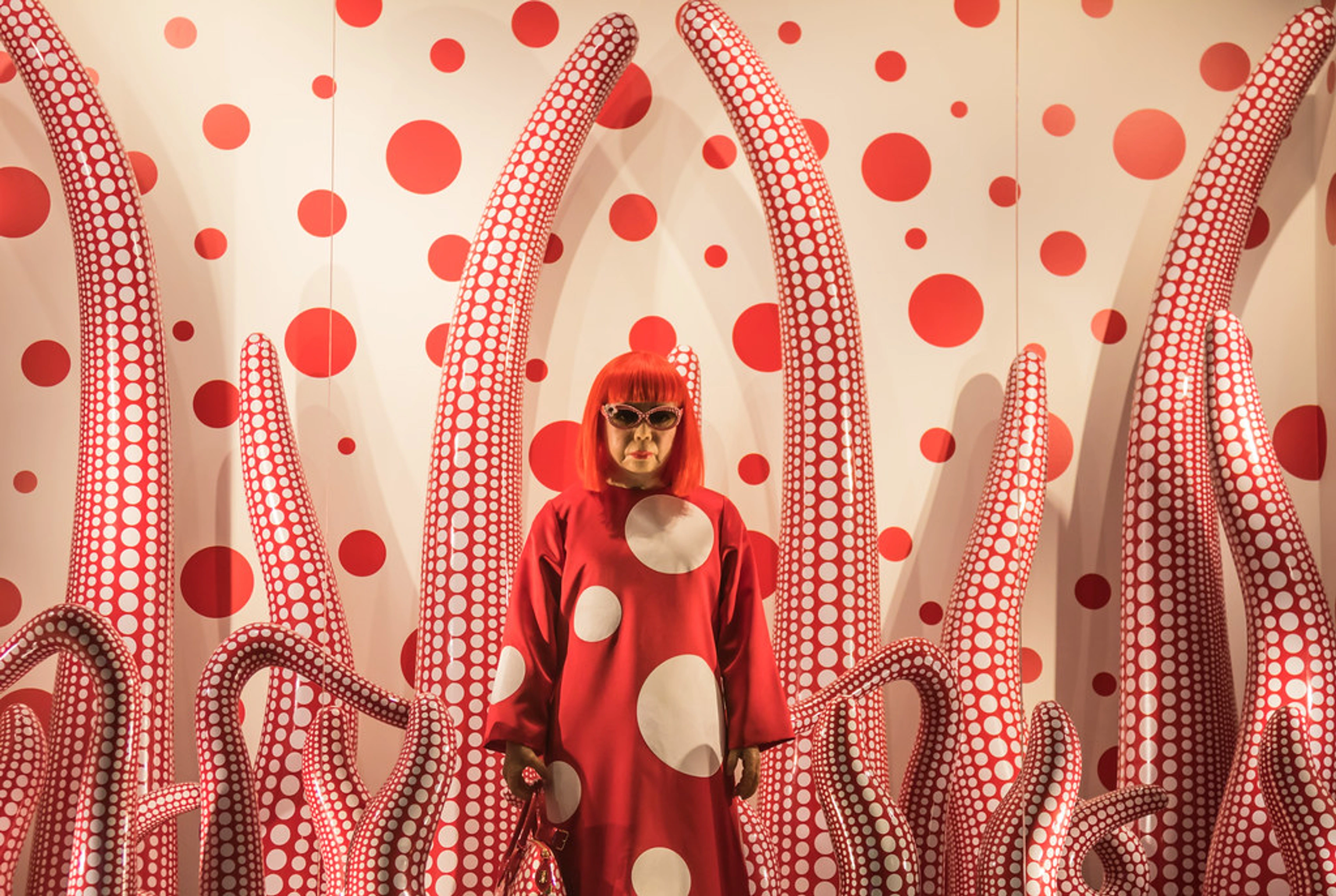Yayoi Kusama, een Japanse kunstenares, staat in een rode polkadot jurk en zonnebril te midden van haar iconische rood-witte polkadot tentakelsculpturen en een bijpassende polkadot muur.