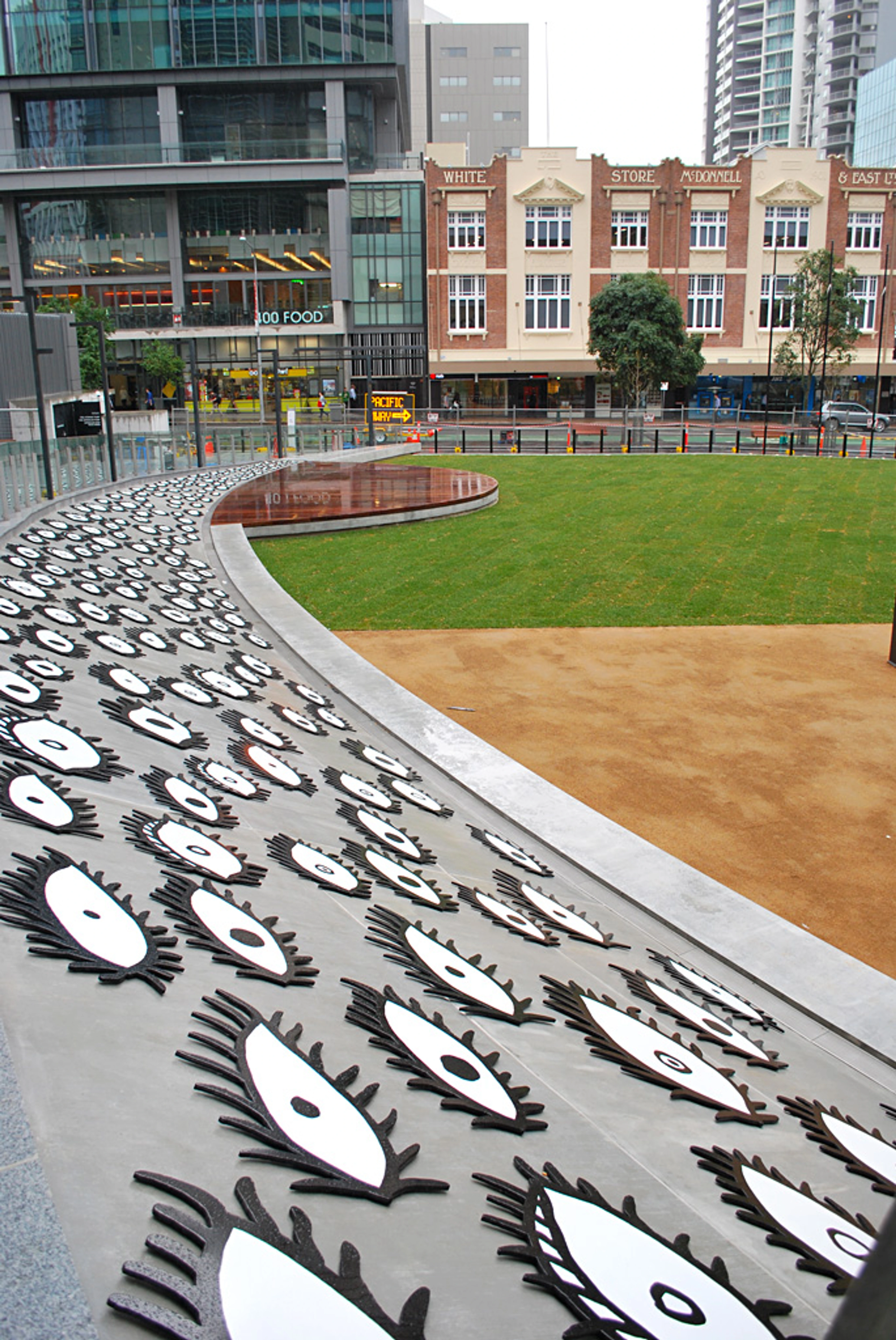 Openbare kunstinstallatie 'Eyes Are Singing Out' van Yayoi Kusama, met talloze zwart-witte oogvormige sculpturen op een gebogen grijs pad, naast een groen gazon en stedelijke gebouwen.