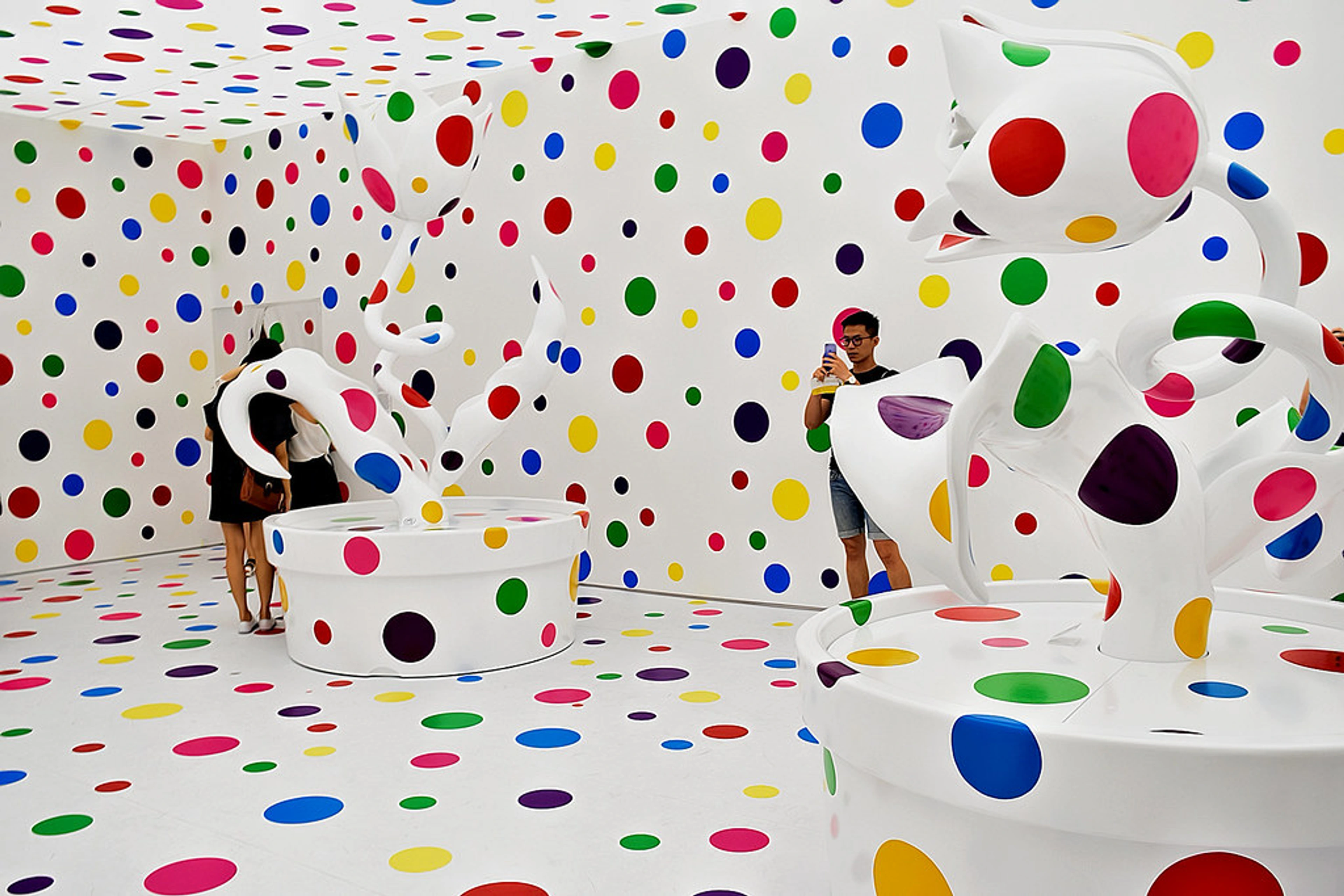 Yayoi Kusama's 'With All My Love for the Tulips, I Pray Forever' installation. A white room with colorful polka dots on walls, floor, and large plant sculptures, with visitors.