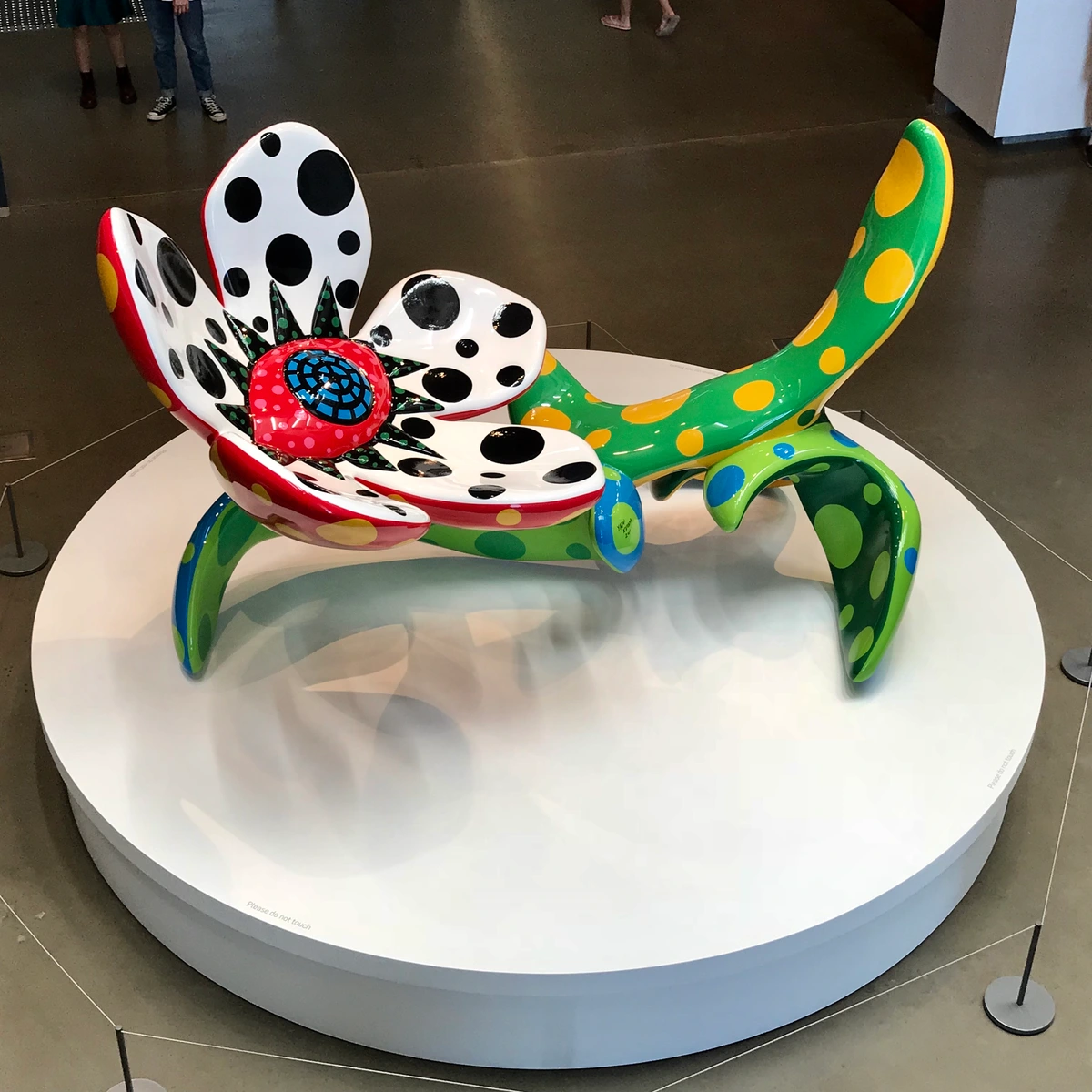 A vibrant, polka-dotted flower sculpture by famous artist Yayoi Kusama, featuring red, green, yellow, and blue colors, displayed on a white circular platform in a museum setting.
