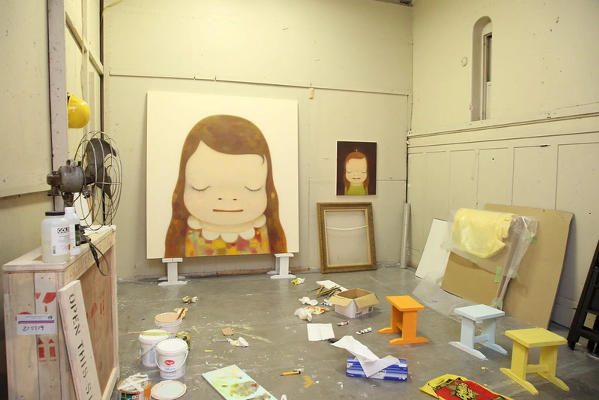 Interior of Yoshitomo Nara's art studio with a large painting of a girl with closed eyes, smaller artworks, paint supplies, and colorful stools.