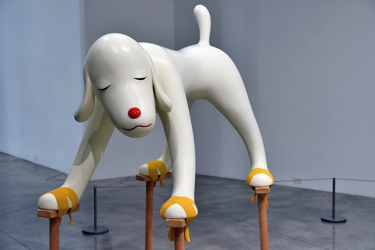 White sculpture by Yoshitomo Nara of a dog with closed eyes and a red nose, standing on stilts with yellow wrappings.