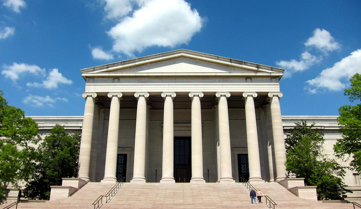 Frontal view of the National Gallery of Art's West Building in Washington, D.C., featuring its neoclassical architecture, columns, and grand staircase under a blue sky with clouds.