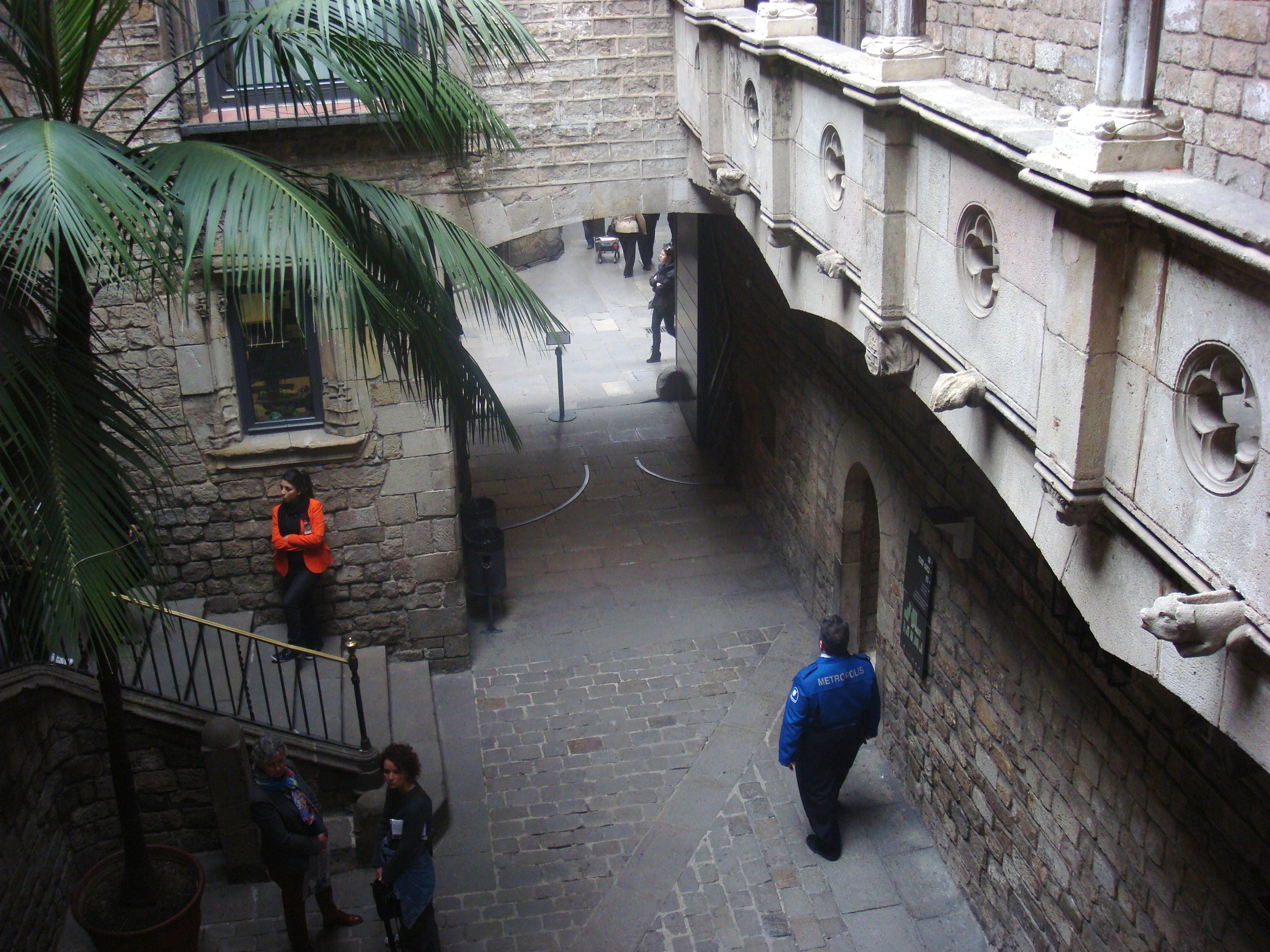 View of the ornate Pont del Bisbe (Bishop's Bridge) in Barcelona's Gothic Quarter, spanning a stone courtyard with a large palm tree and people.