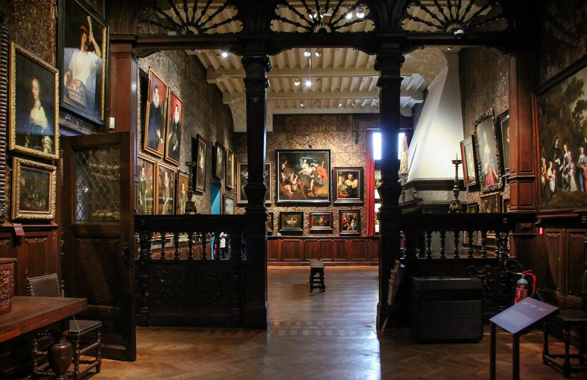 Interior view of a gallery room in the Mauritshuis museum, The Hague, featuring numerous Dutch Golden Age paintings on patterned walls, ornate dark wood paneling, and a parquet floor.