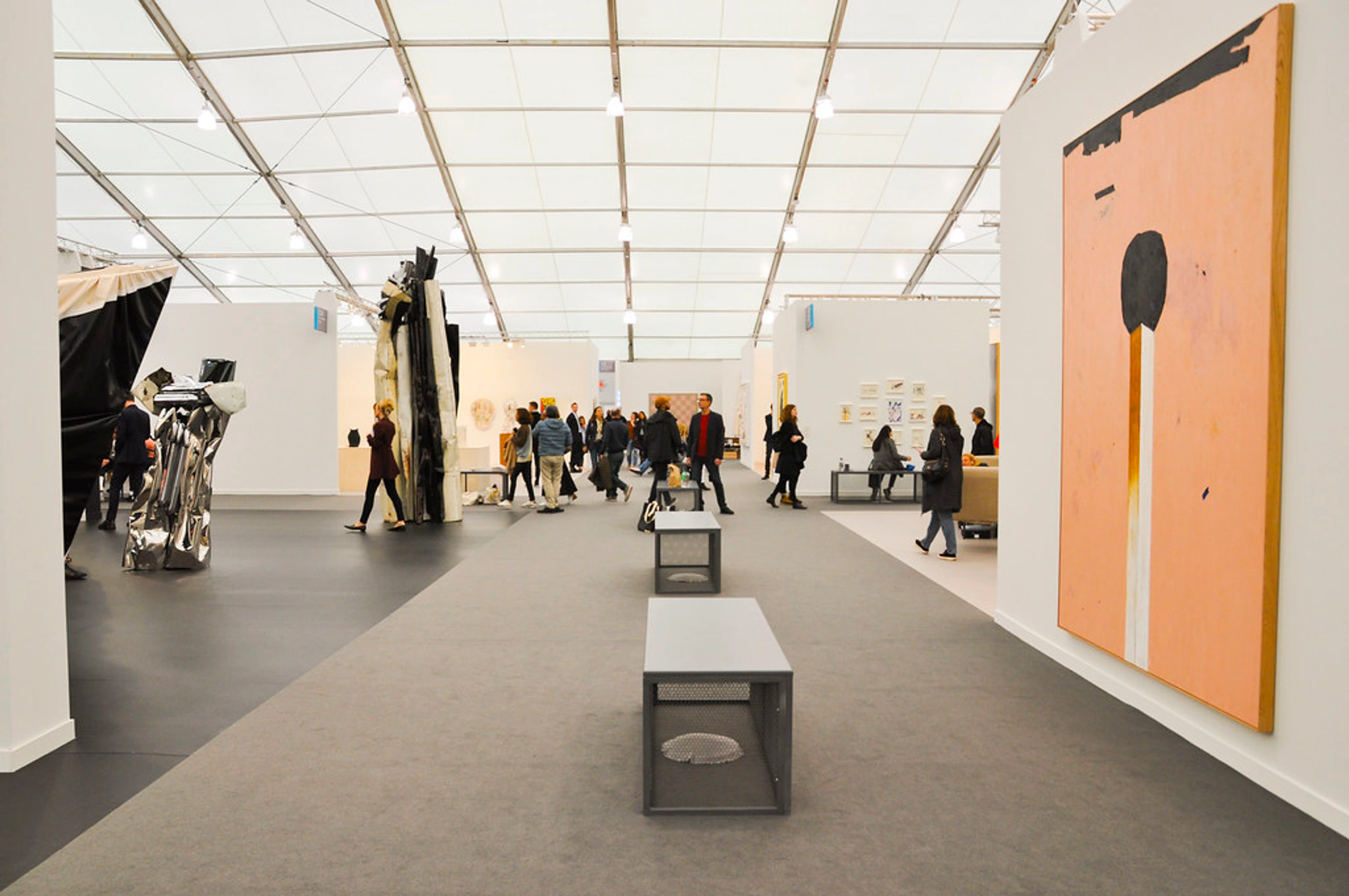 Interior view of a bustling contemporary art fair with numerous visitors browsing various artworks, including large abstract sculptures and paintings, under a white tented ceiling.