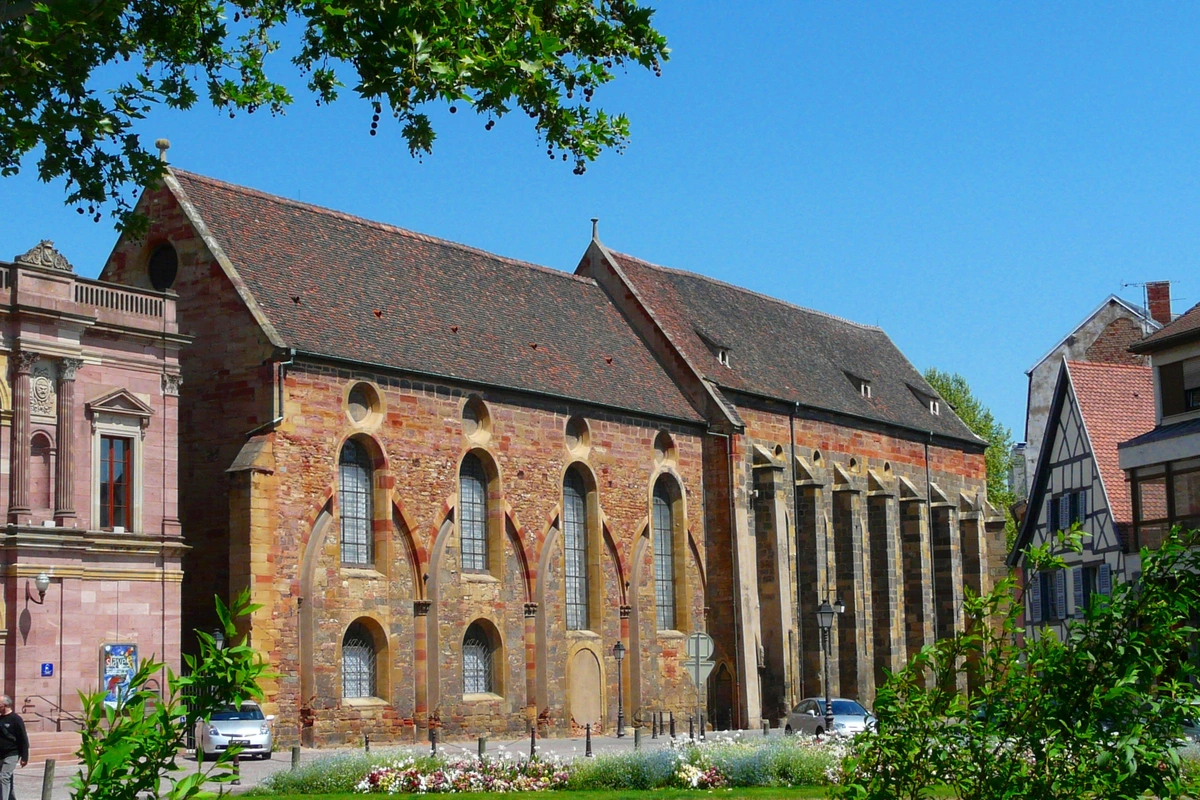 The historic Koïfhus (Old Customs House) in Colmar, France, a medieval building with a red-tiled roof and stone walls, under a clear blue sky. Traditional half-timbered houses and classical architecture are visible nearby.