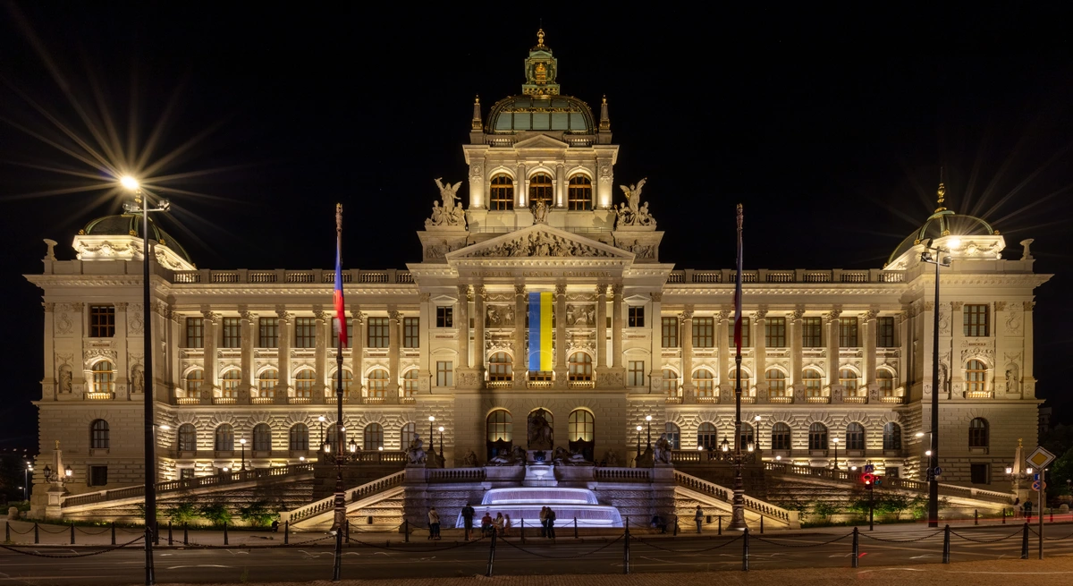The grand National Museum in Prague, Czech Republic, illuminated at night, featuring the Czech and Ukrainian flags and a glowing fountain.