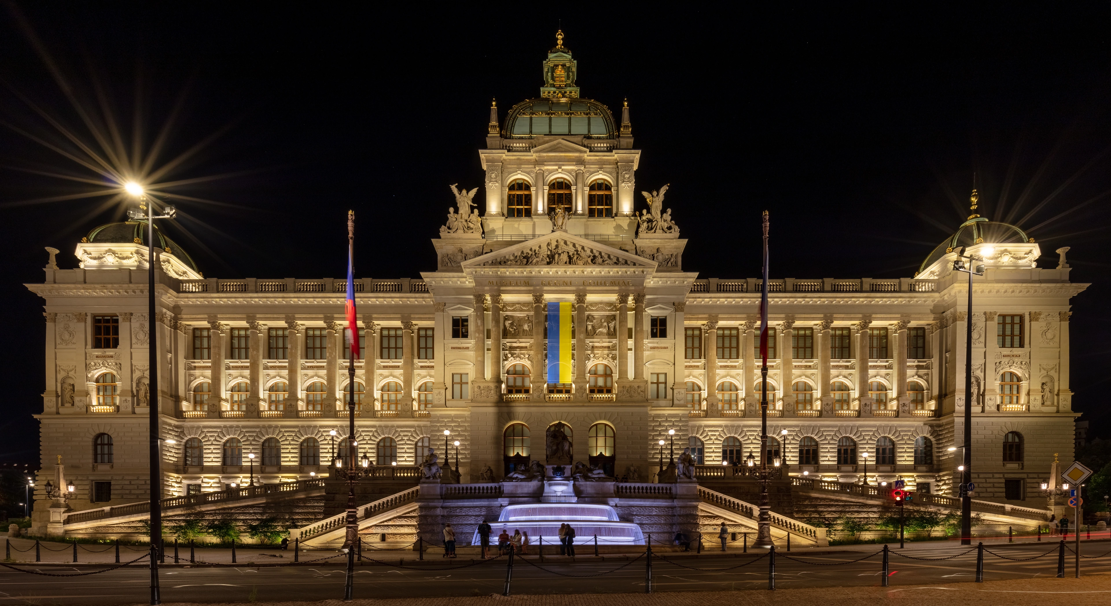 The grand National Museum in Prague, Czech Republic, illuminated at night, featuring the Czech and Ukrainian flags and a glowing fountain.