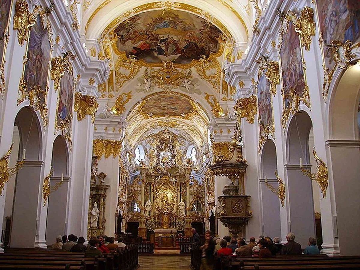 Intricate interior of a baroque church with white walls, ornate gold decorations, ceiling frescoes, and people seated in pews.