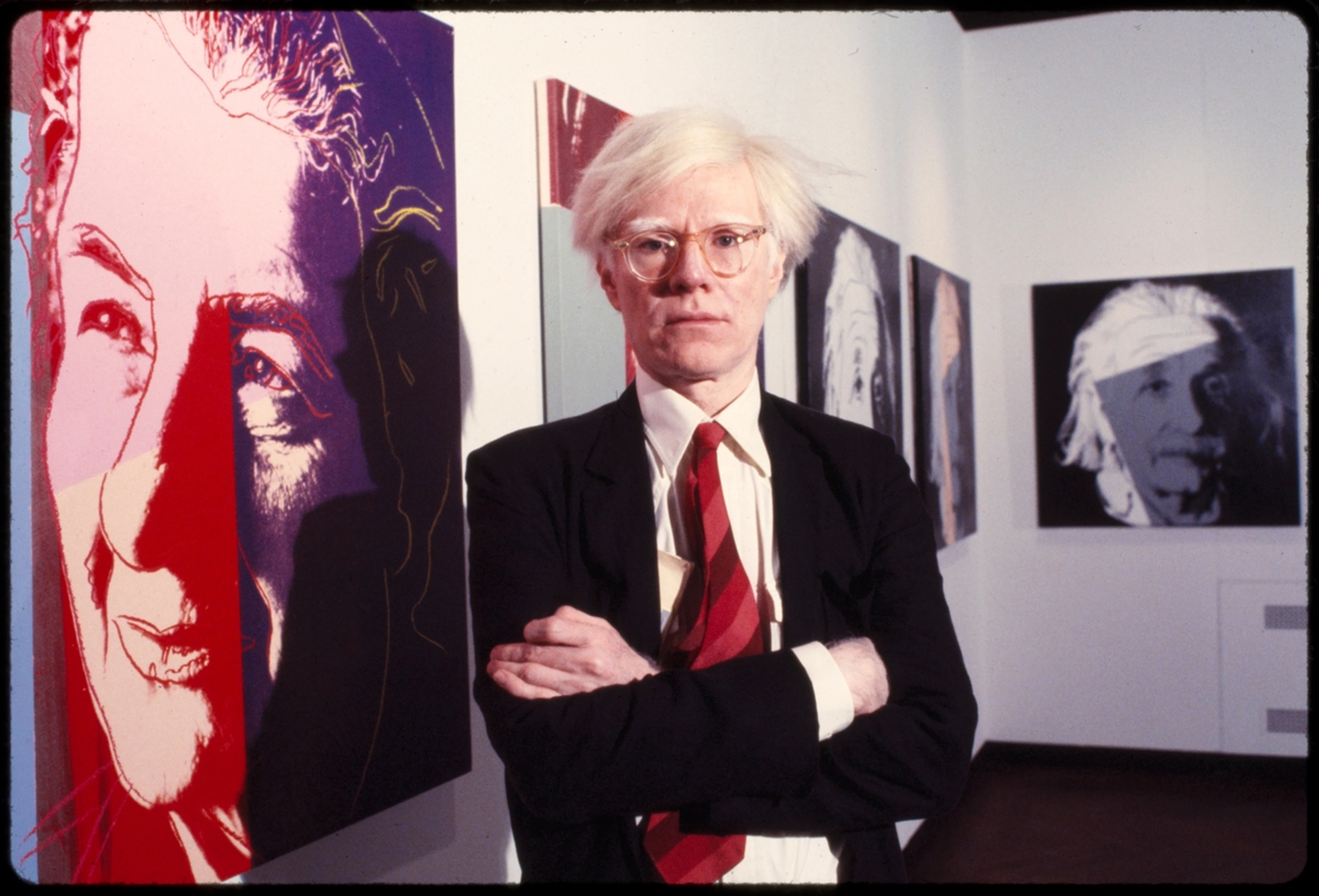 Color photograph of Andy Warhol with his arms crossed, standing in front of several of his self-portrait screen prints in varying colors.