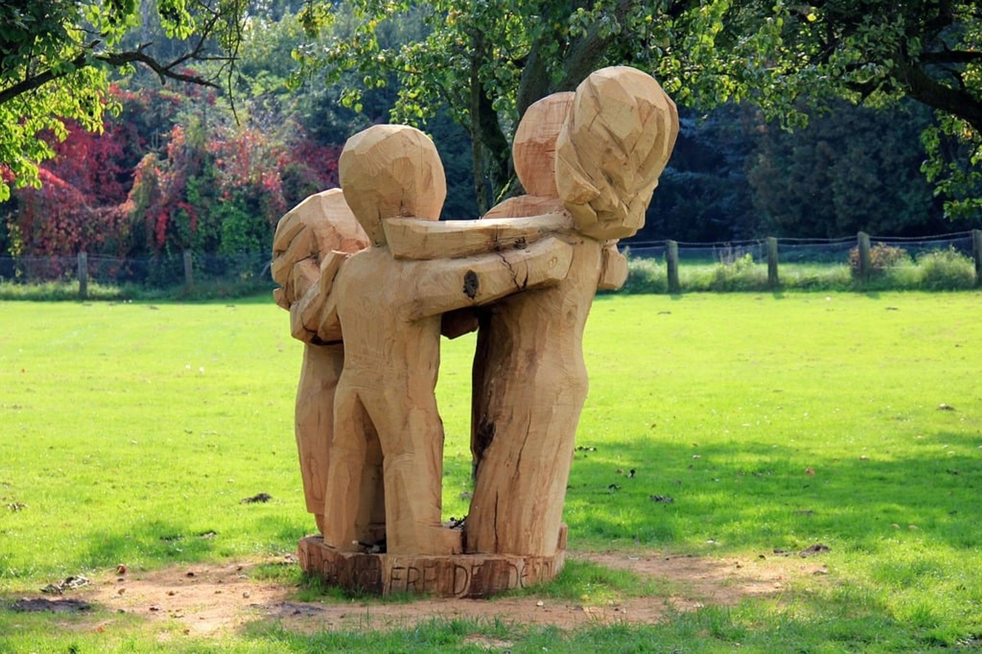 A rustic wooden sculpture depicting four stylized figures embracing in a circle, situated on a grassy area surrounded by trees, some with autumn foliage. The sculpture appears to be carved from a tree trunk and rests on a base of wood chips.