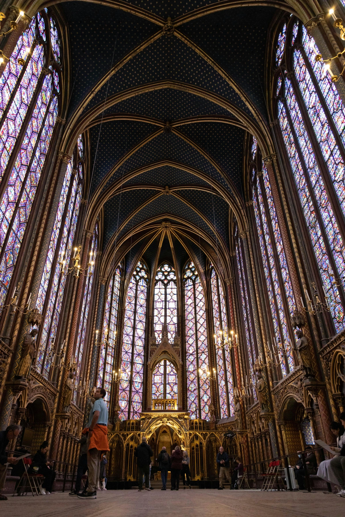 Interior view of Sainte-Chapelle in Paris, showcasing its tall stained glass windows filling the space with colorful light and intricate Gothic architecture. Visitors are visible in the foreground.