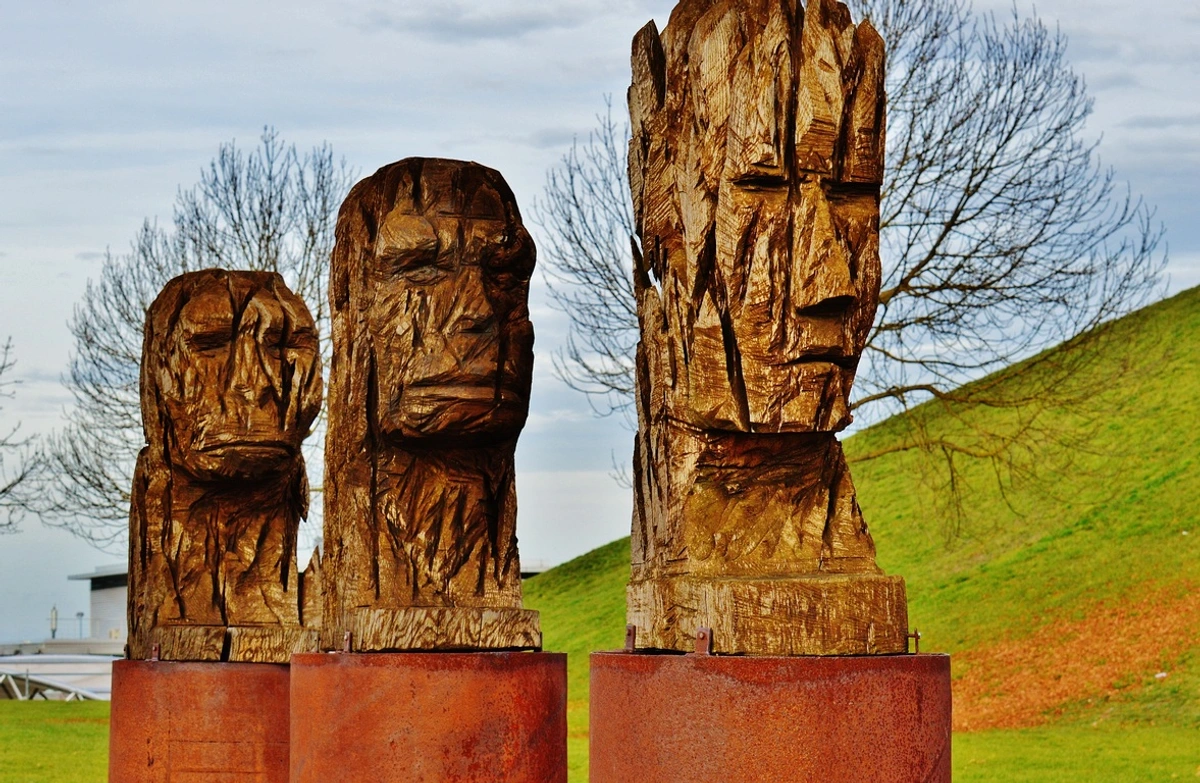 Three tall, roughly carved wooden sculptures of human heads with expressive faces, mounted on reddish-brown pedestals outdoors against a backdrop of a grassy hill and trees under an overcast sky.