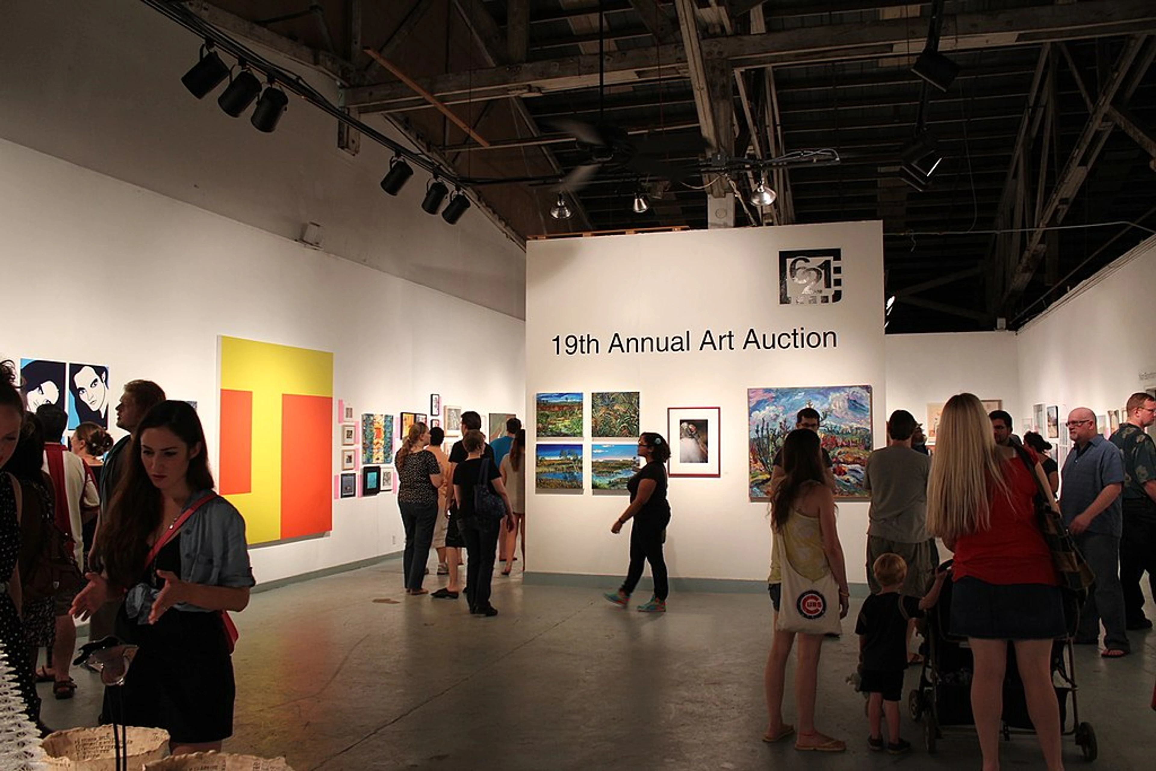 Interior of a lively art auction with numerous attendees viewing a variety of artworks displayed on the white walls. A sign reads "19th Annual Art Auction". Track lighting illuminates the space.