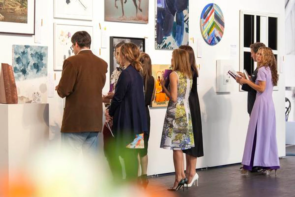 Several people are gathered in a brightly lit art exhibition, attentively looking at various paintings and a sculpture displayed on a white wall.