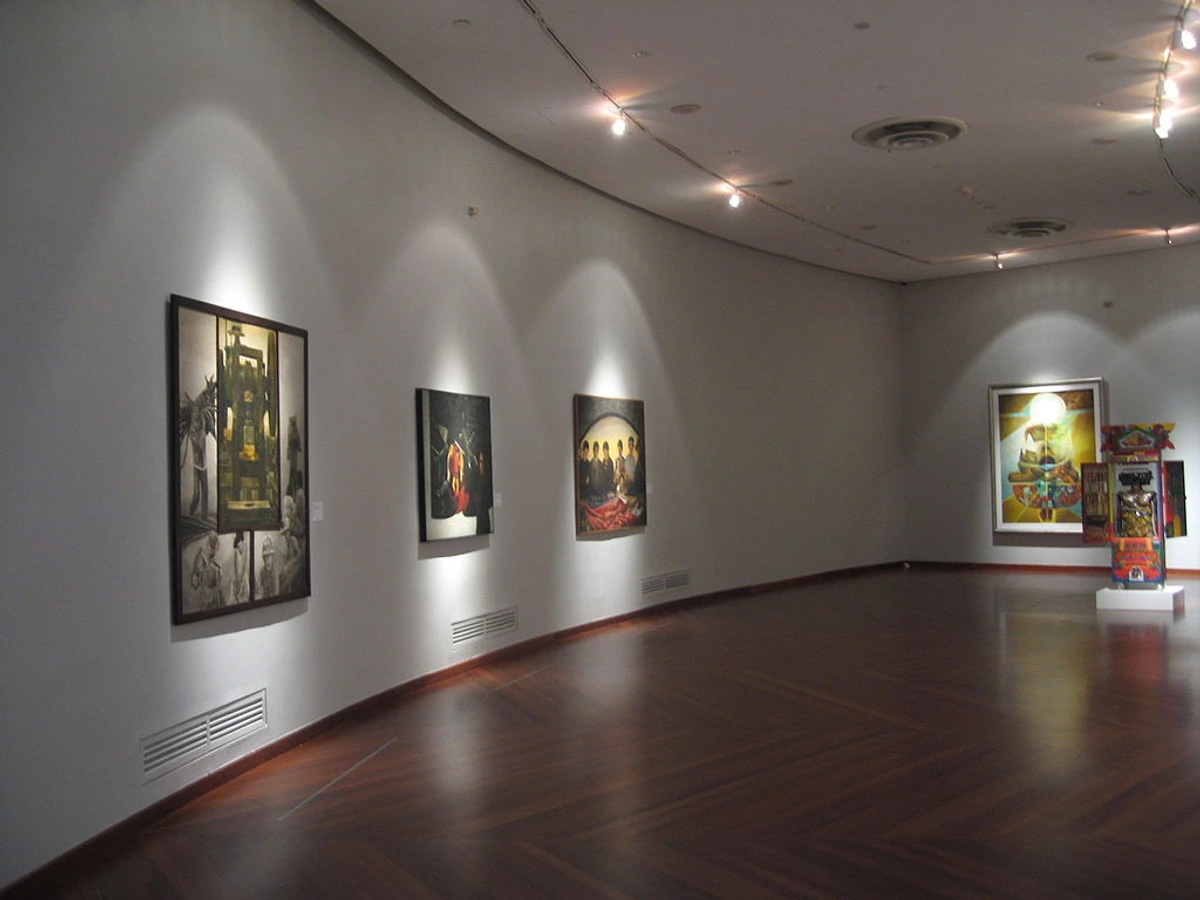 Curved museum gallery with dark wooden floors, white walls illuminated by spotlights, and several framed paintings on display.