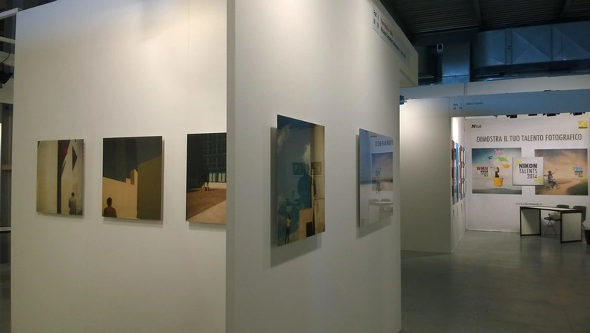 Uitzicht op een witte gang bij een kunsttentoonstelling, met minimalistische schilderijen met figuren en architectonische elementen.