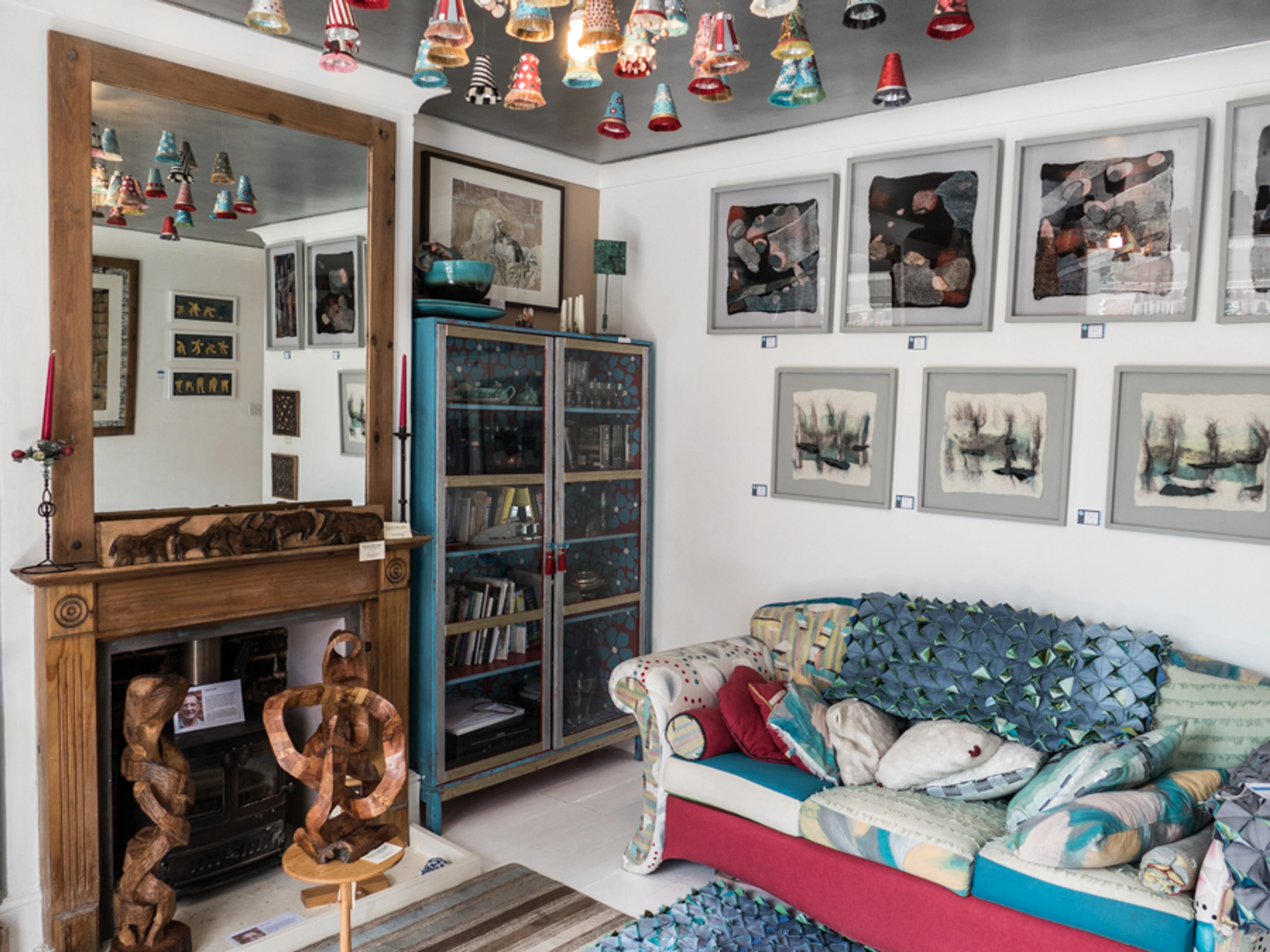 Eclectic living space or gallery with fireplace, mirror, sculptures, blue cabinet, hanging glass ornaments, patterned sofa, and abstract art.