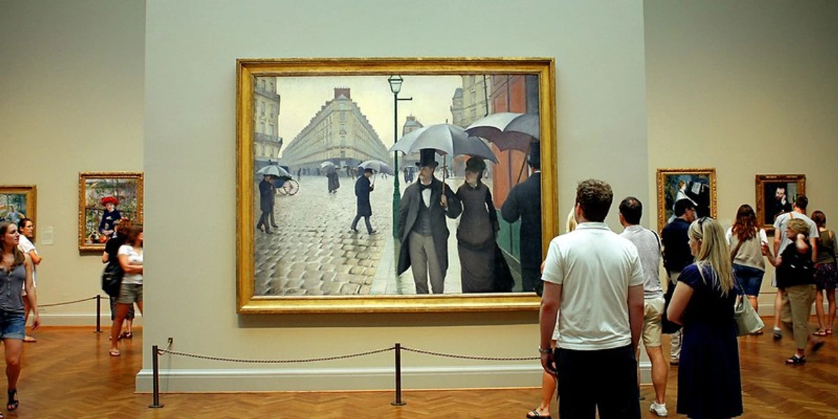 Museumbezoekers kijken naar Gustave Caillebotte's "Paris Street; Rainy Day" in een galerieomgeving.