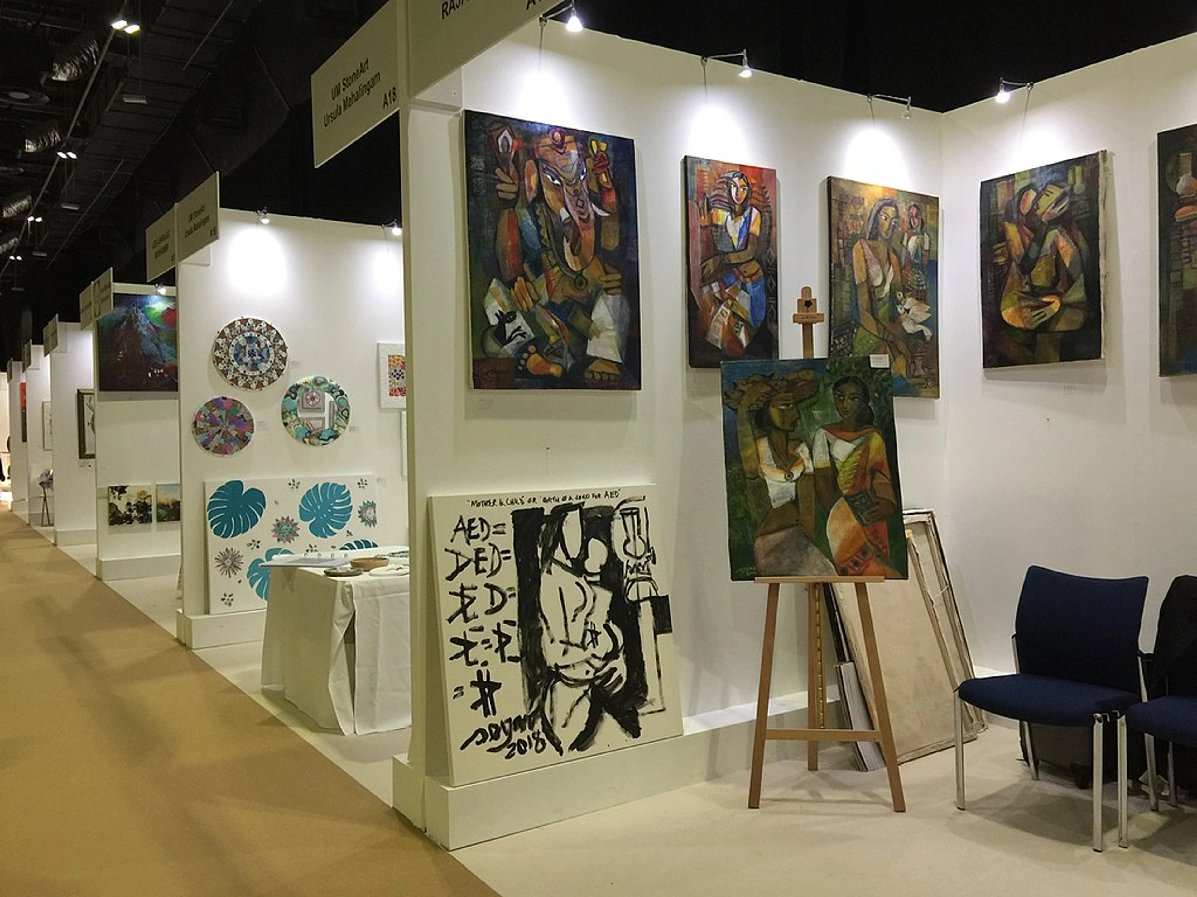 View of an art fair booth with various colorful paintings displayed on the white walls and one painting on a wooden easel.