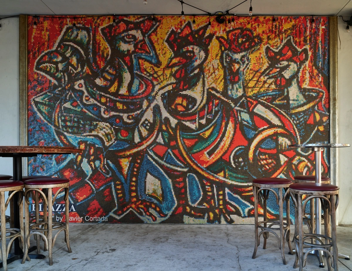 Colorful mosaic mural by Avier Cortada featuring abstract figures, possibly birds, with tables and stools in front of it.