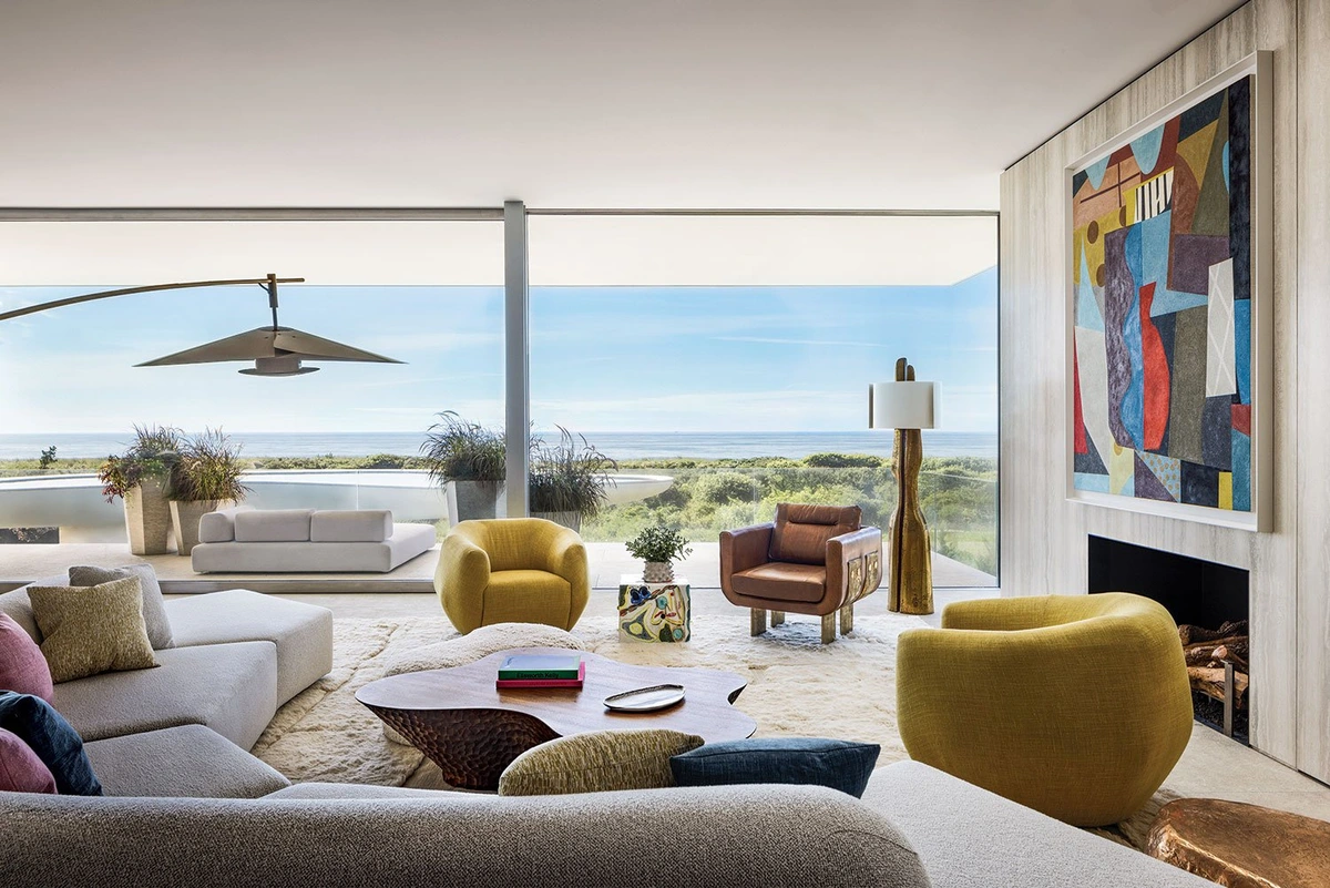 Lichte en luchtige moderne woonkamer met grote ramen met uitzicht op de oceaan, met een hoekbank, fauteuils, een unieke salontafel en een abstract schilderij boven een open haard.