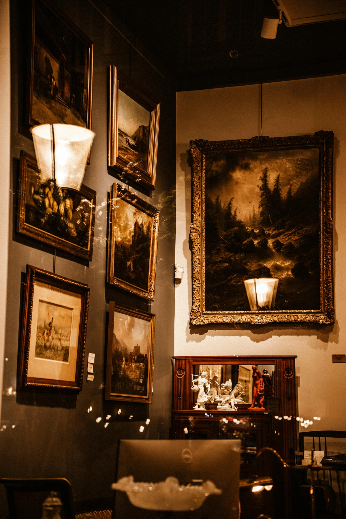 Interieur van een schemerige kunstgalerie met verschillende ingelijste landschapsschilderijen aan de muur en een reflecterend oppervlak onder een groot schilderij.