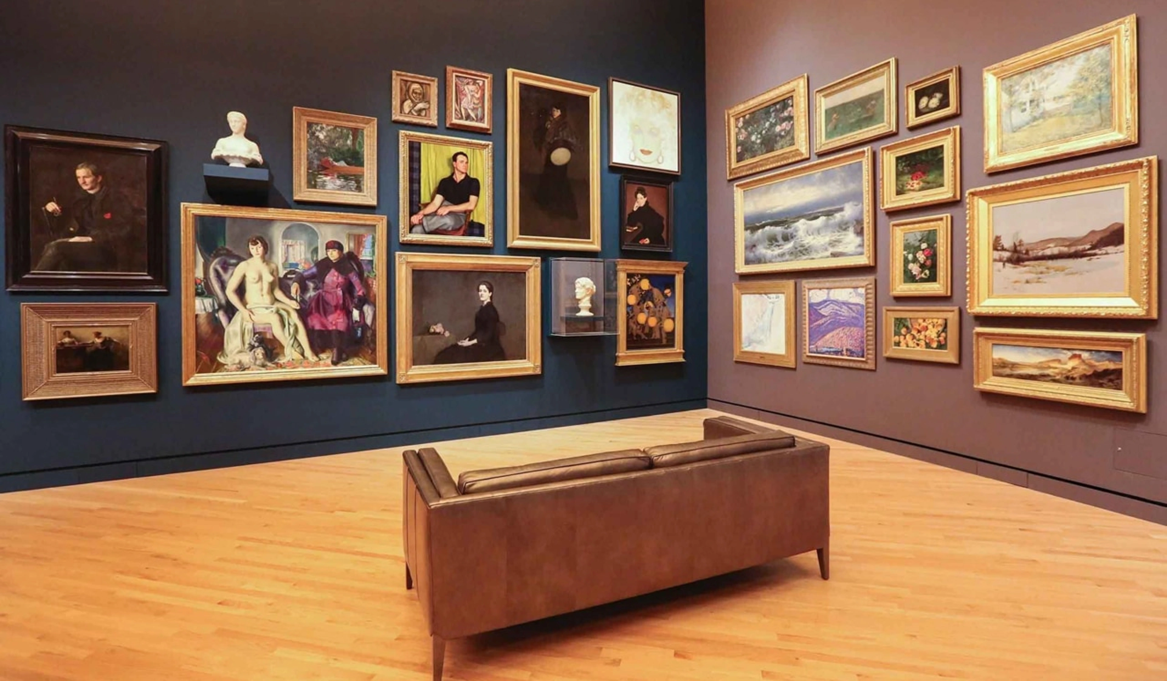 Art gallery interior with numerous framed paintings and a white bust displayed on dark blue and brown walls, and a brown leather sofa in the foreground.