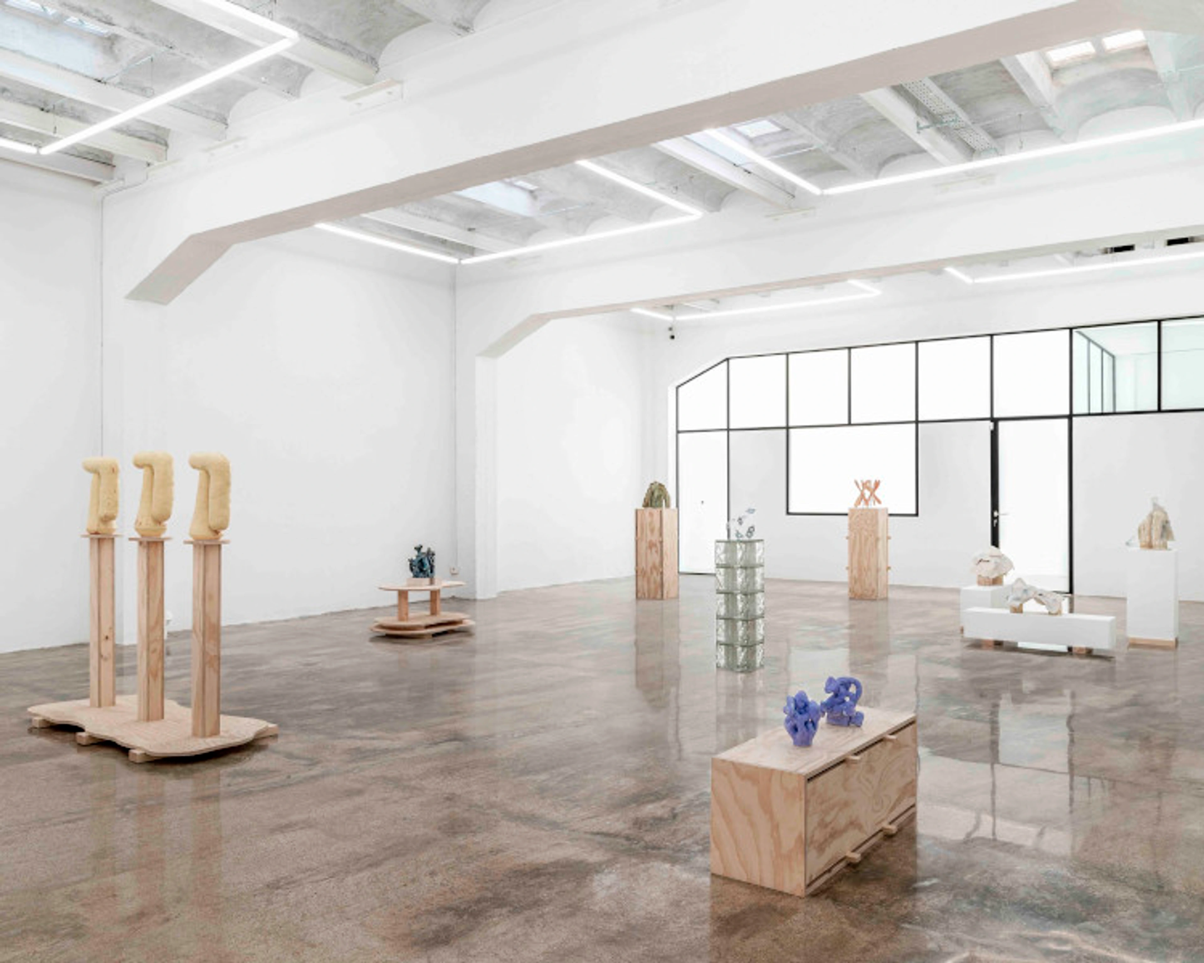 Bright and spacious modern art gallery with various contemporary sculptures displayed on pedestals and directly on the polished concrete floor. Large windows provide natural light.
