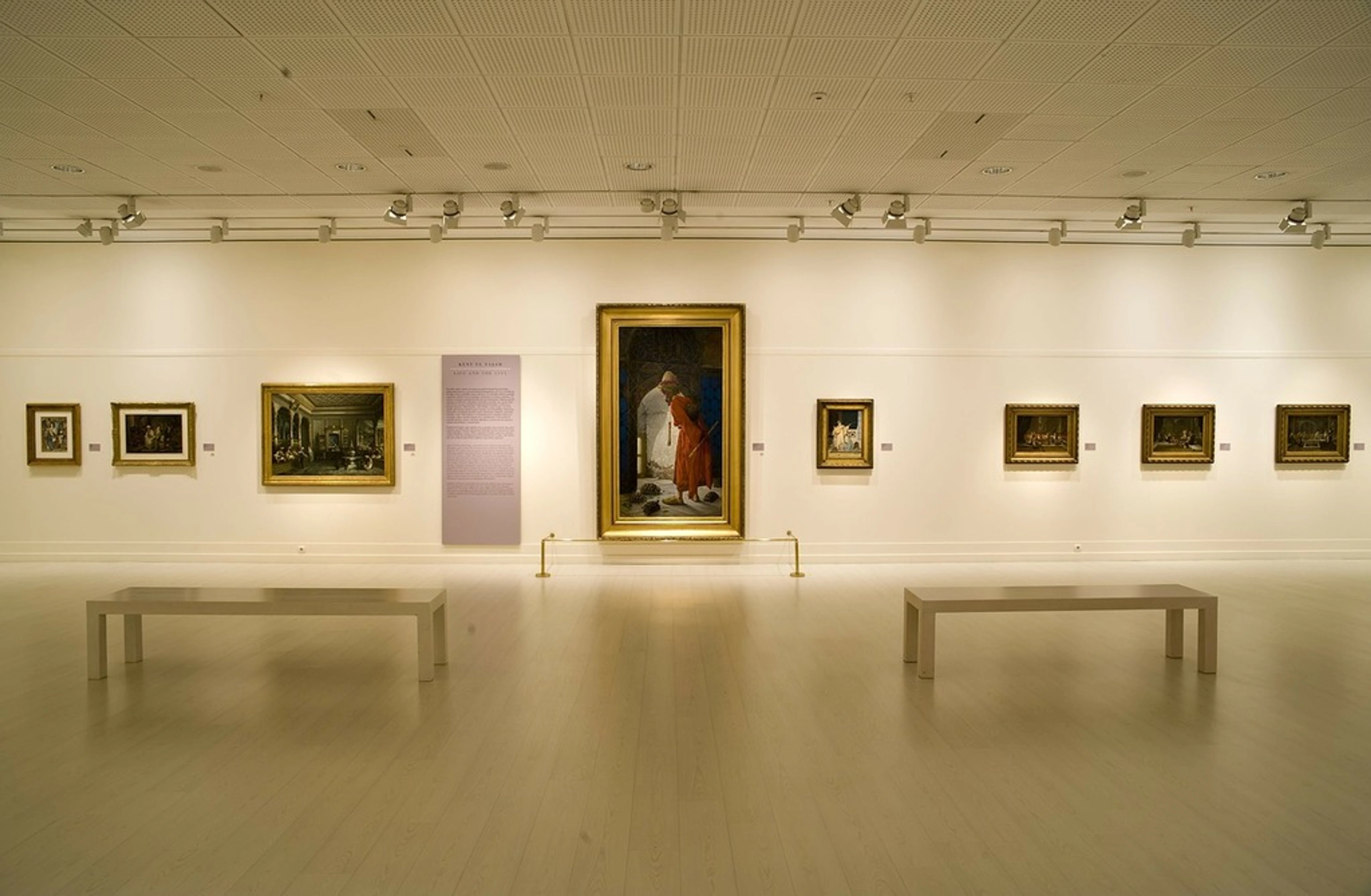 Wide view of a museum gallery with several framed paintings on a white wall, two benches in the foreground, and track lighting above.