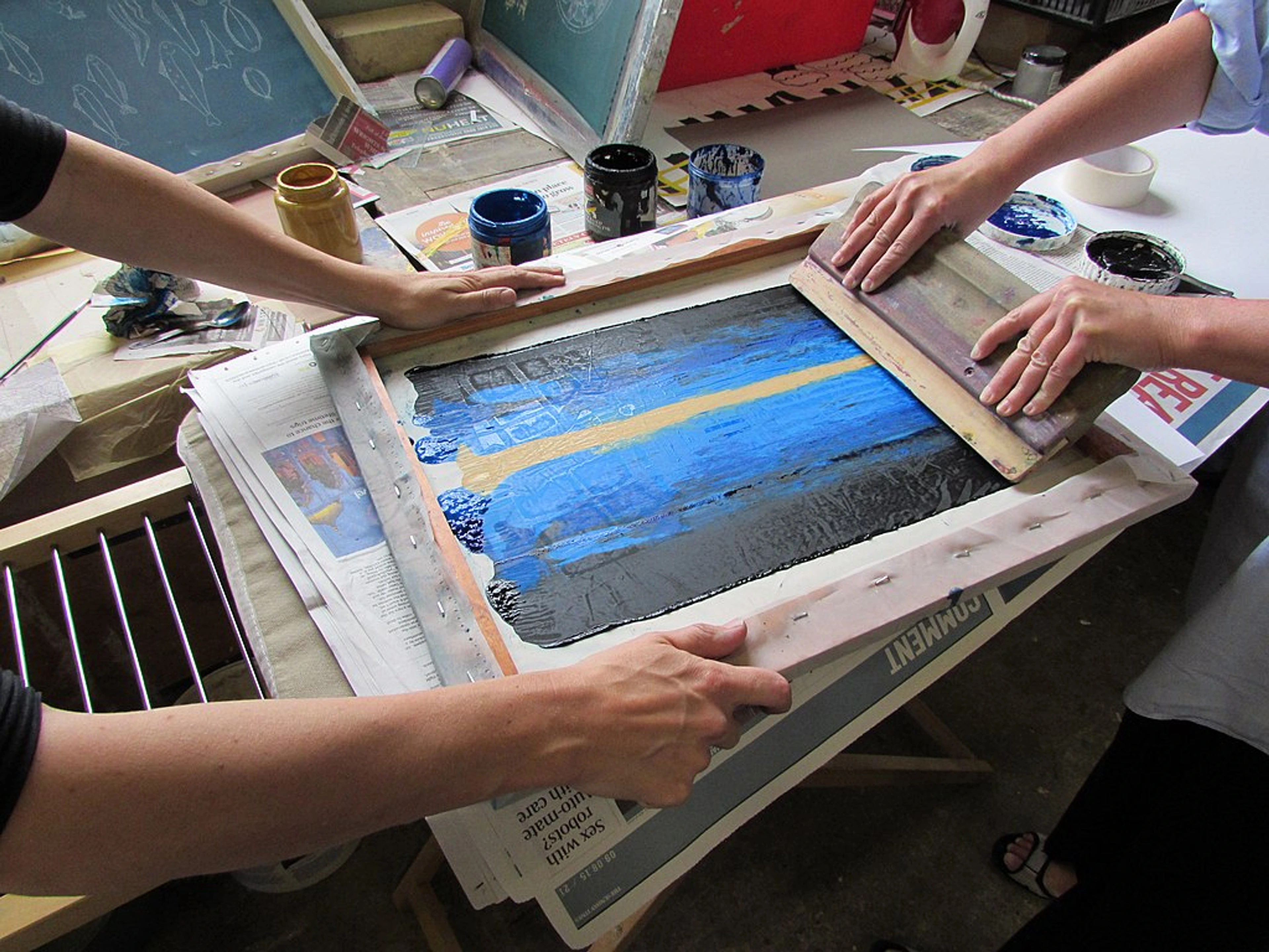 Overhead view of three pairs of hands engaged in the screen printing process. Ink is being spread across a screen, likely in a workshop or art studio setting with various supplies and newspapers visible on the work surface.