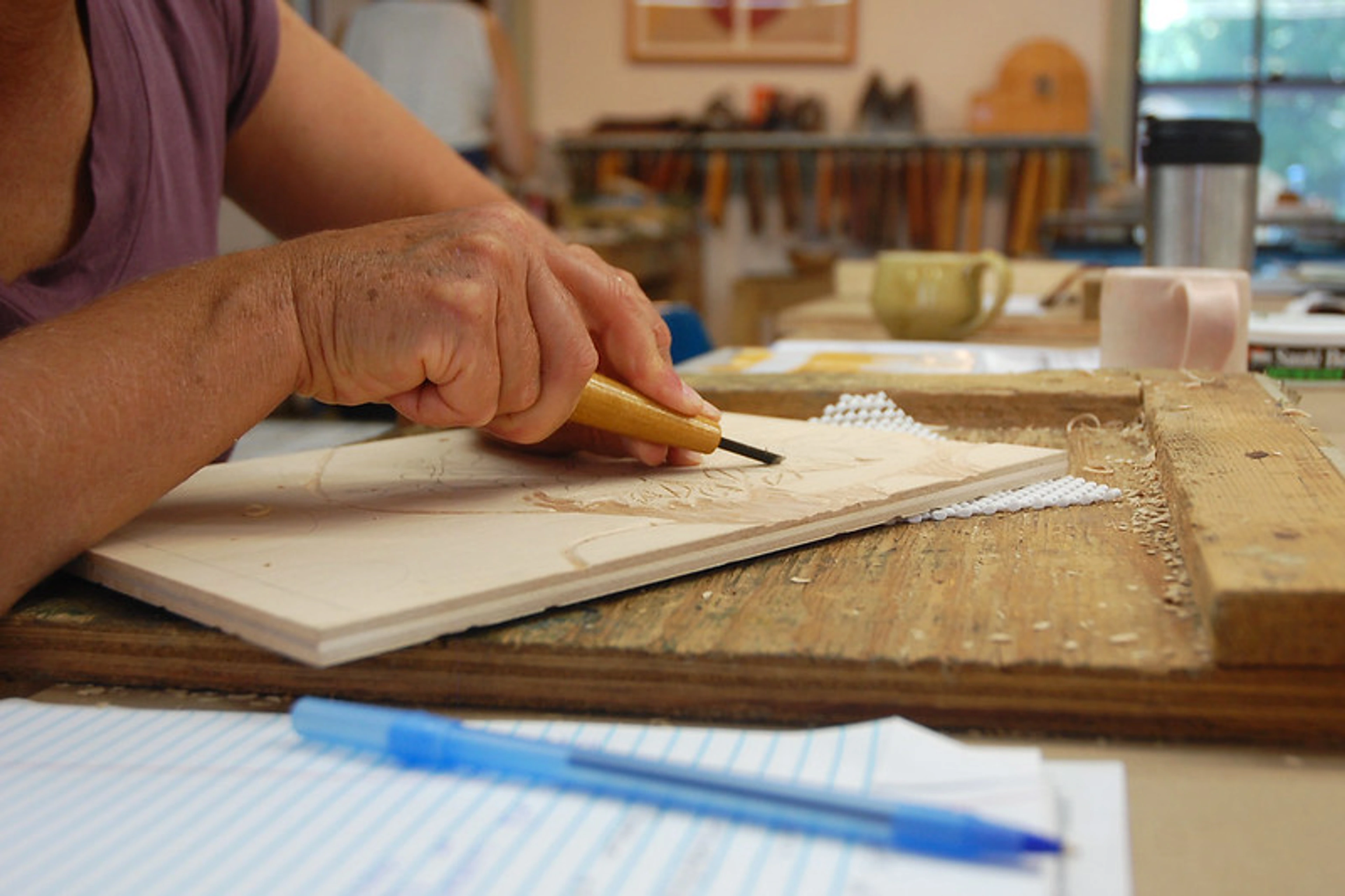 Close-up of hands using a small carving tool to meticulously cut a design into a light-colored woodblock. Wood shavings accumulate around the carved area.