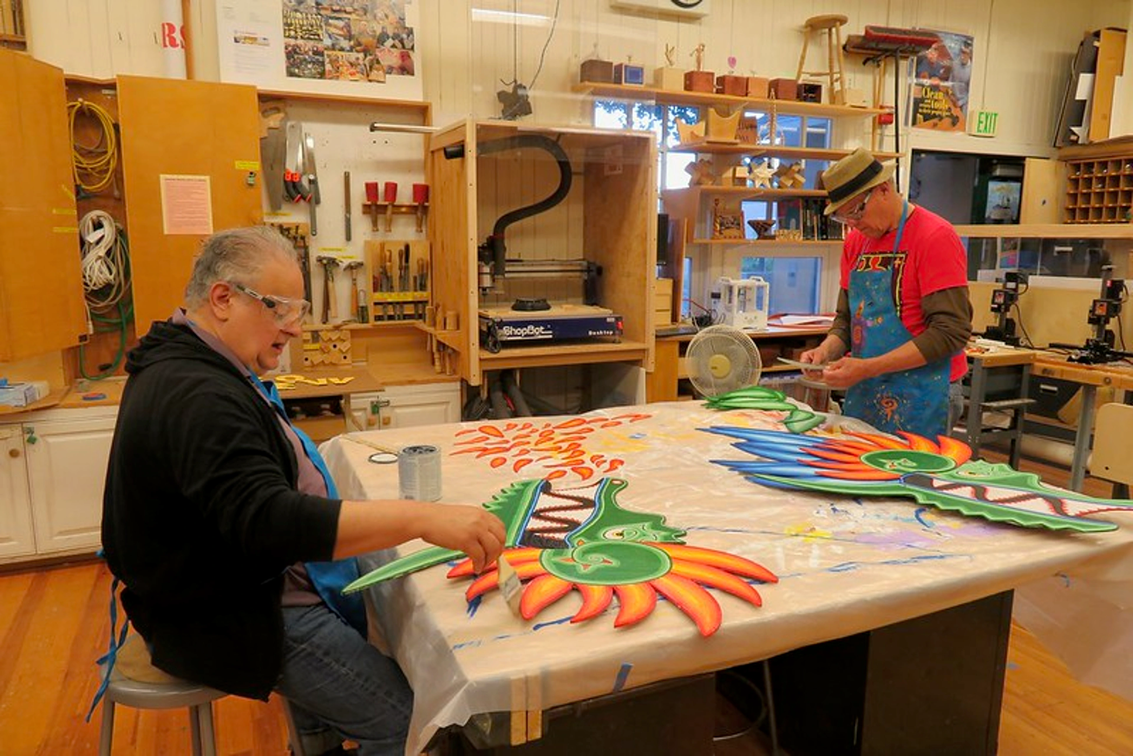 Two artists are working in a cluttered studio space. One seated artist is painting a colorful wooden cutout, while another standing artist is working at a nearby table. Tools, supplies, and finished pieces are visible throughout the workshop.