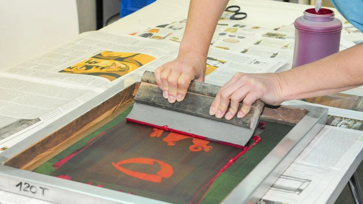 Close-up of hands using a squeegee to push ink through a screen printing frame onto a surface below. A jar of red ink and printed samples on newspaper are visible on the table.