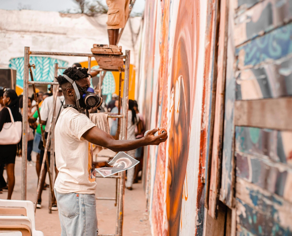 A street artist wearing a respirator mask is spray-painting a large, colorful mural on a wall outdoors, with onlookers and scaffolding nearby.