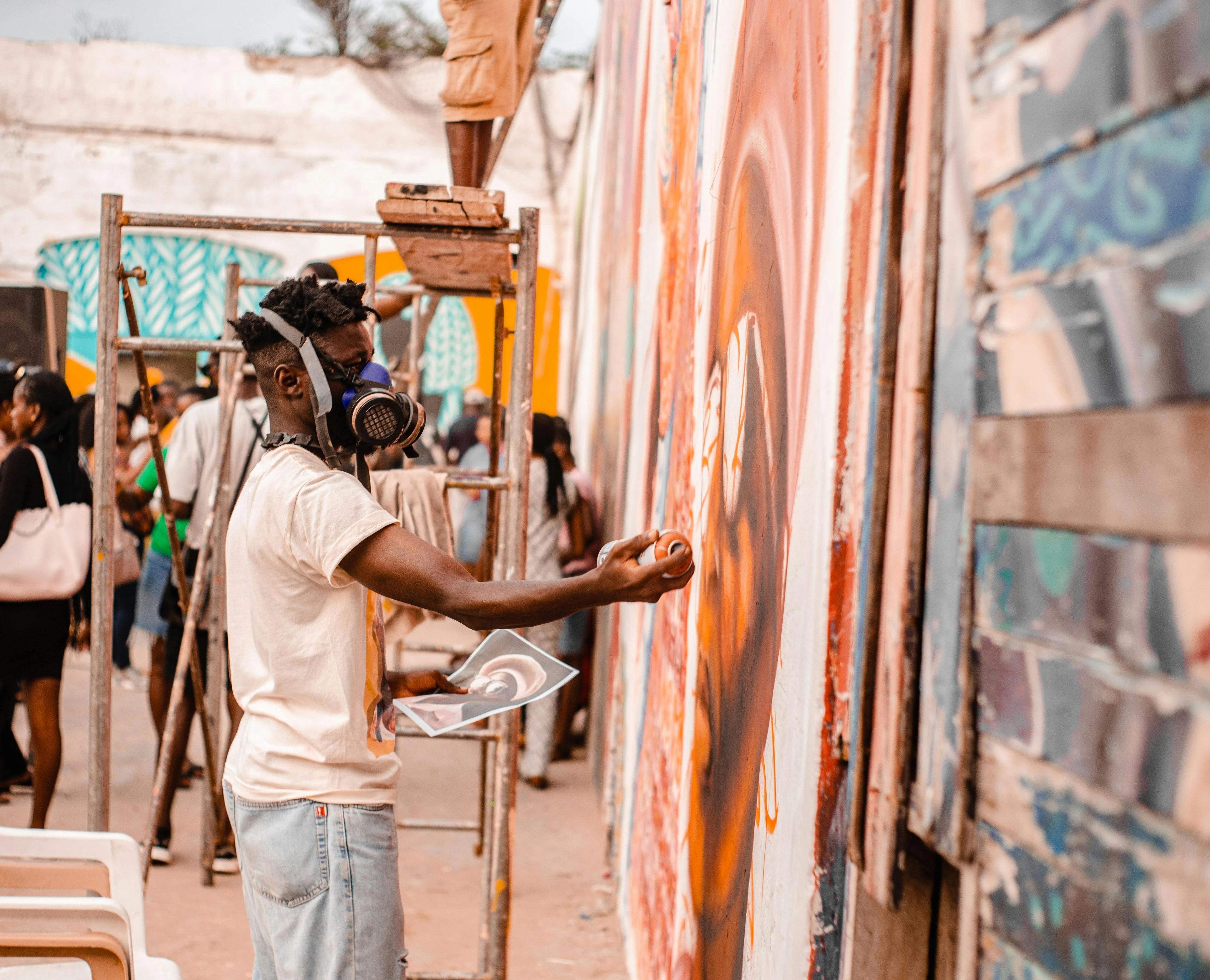 A street artist wearing a respirator mask is spray-painting a large, colorful mural on a wall outdoors, with onlookers and scaffolding nearby.