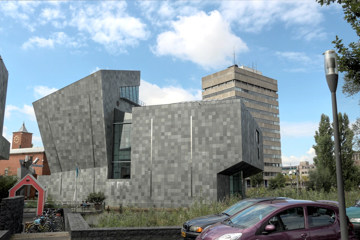The Van Abbemuseum in Eindhoven, Netherlands, a modern building with a grey, geometric facade, under a cloudy sky. A tall building is visible behind it, with cars and bicycles parked in the foreground.
