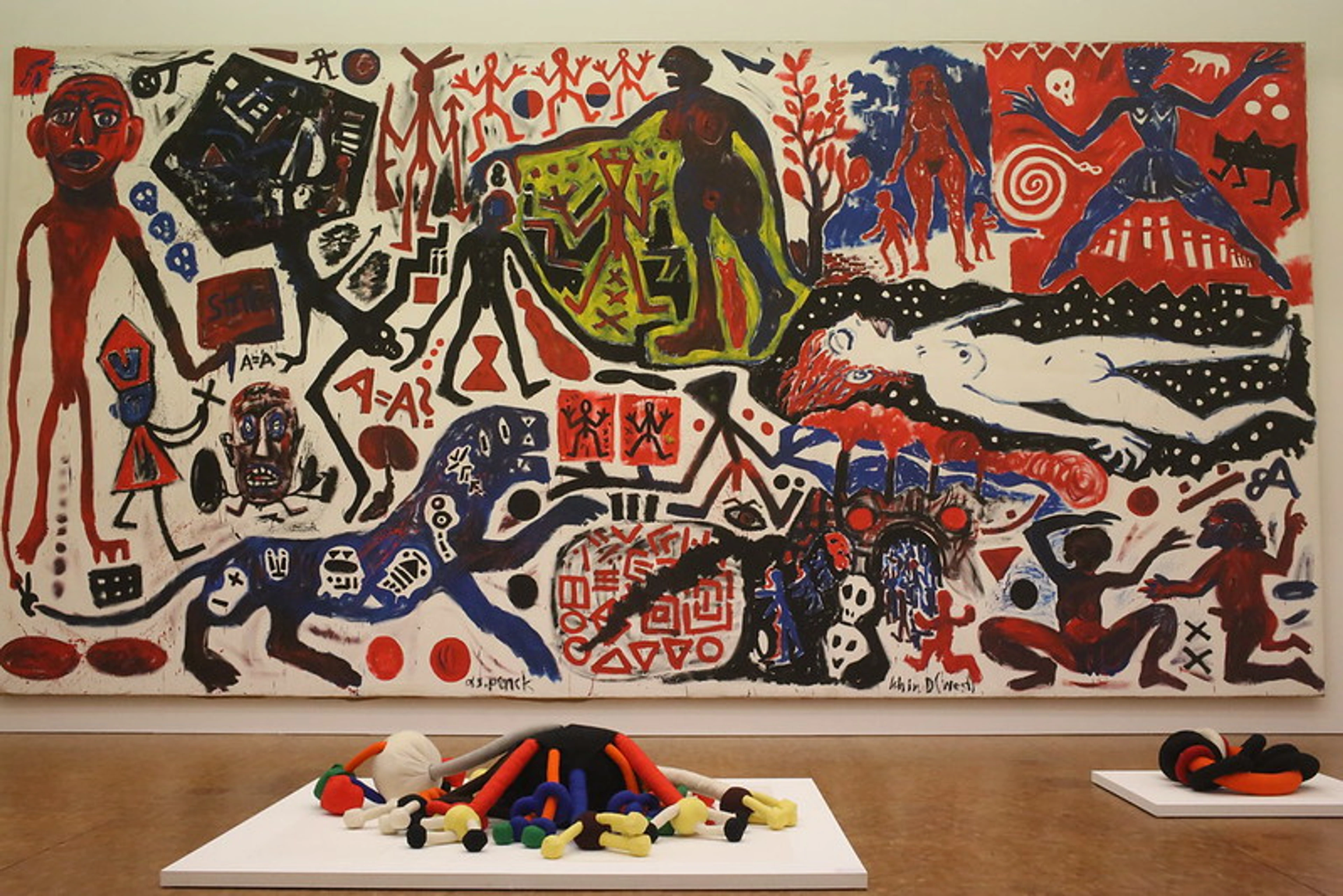 Groot abstract schilderij van A. R. Penck met talrijke figuren en symbolen in rood, zwart, blauw en wit, tentoongesteld boven twee hedendaagse sculpturen.