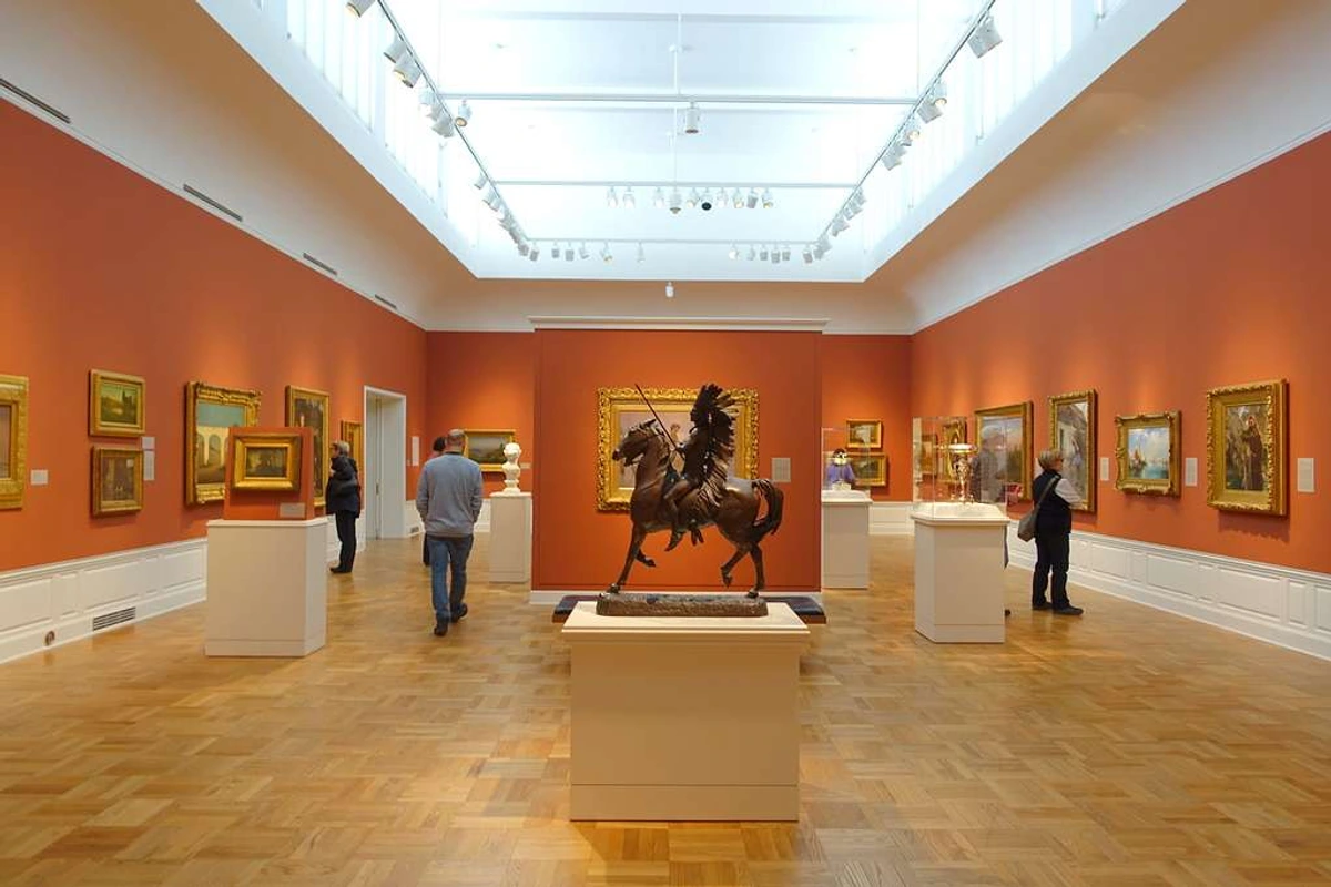 Interieur van een kunstgalerie met schilderijen aan de muren en een bronzen beeld van een inheemse krijger te paard in het midden.